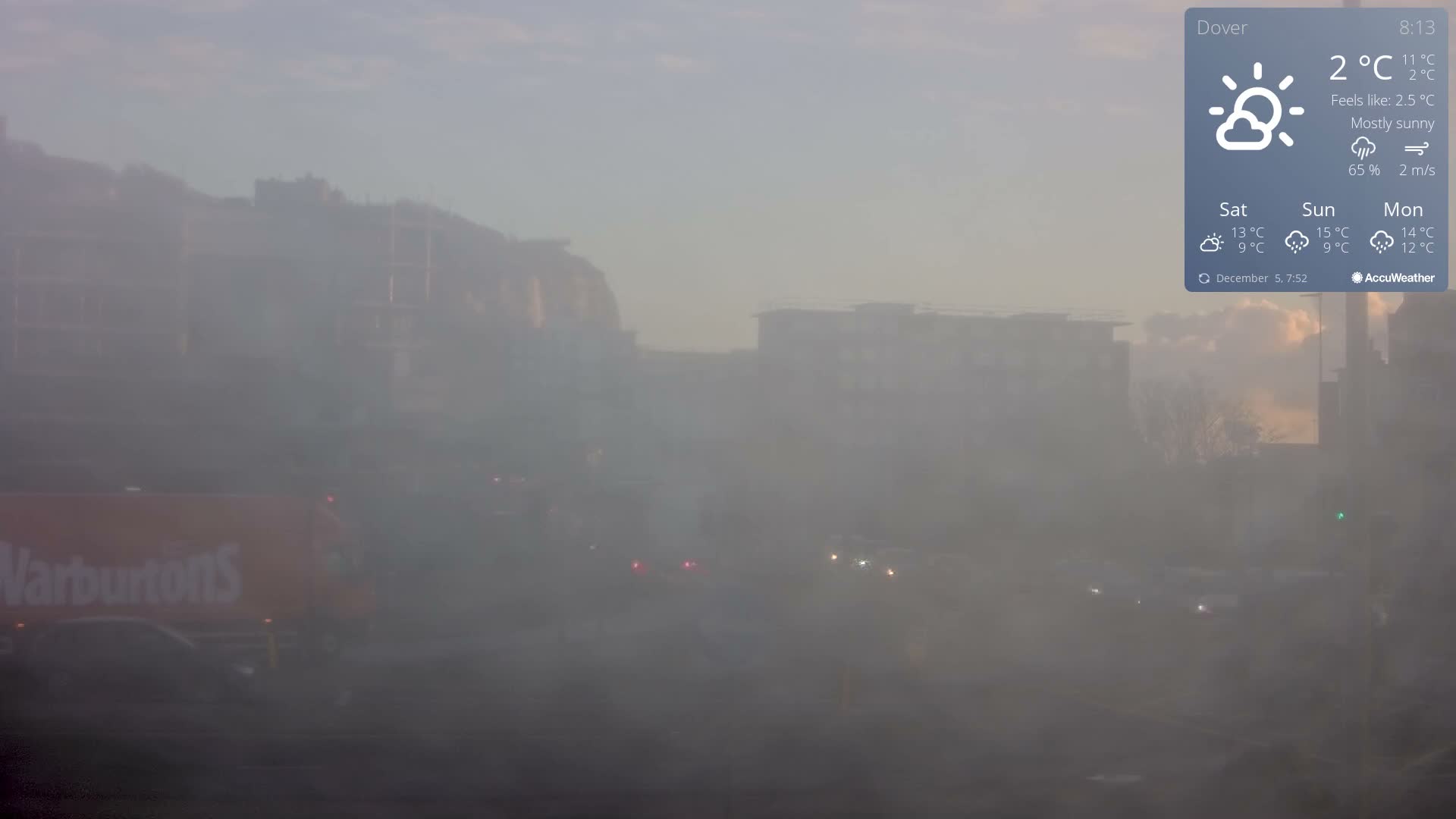 An urban street scene is heavily obscured by dense fog or mist, revealing only the faint outlines of distant buildings and the diffused lights of vehicles on the road.