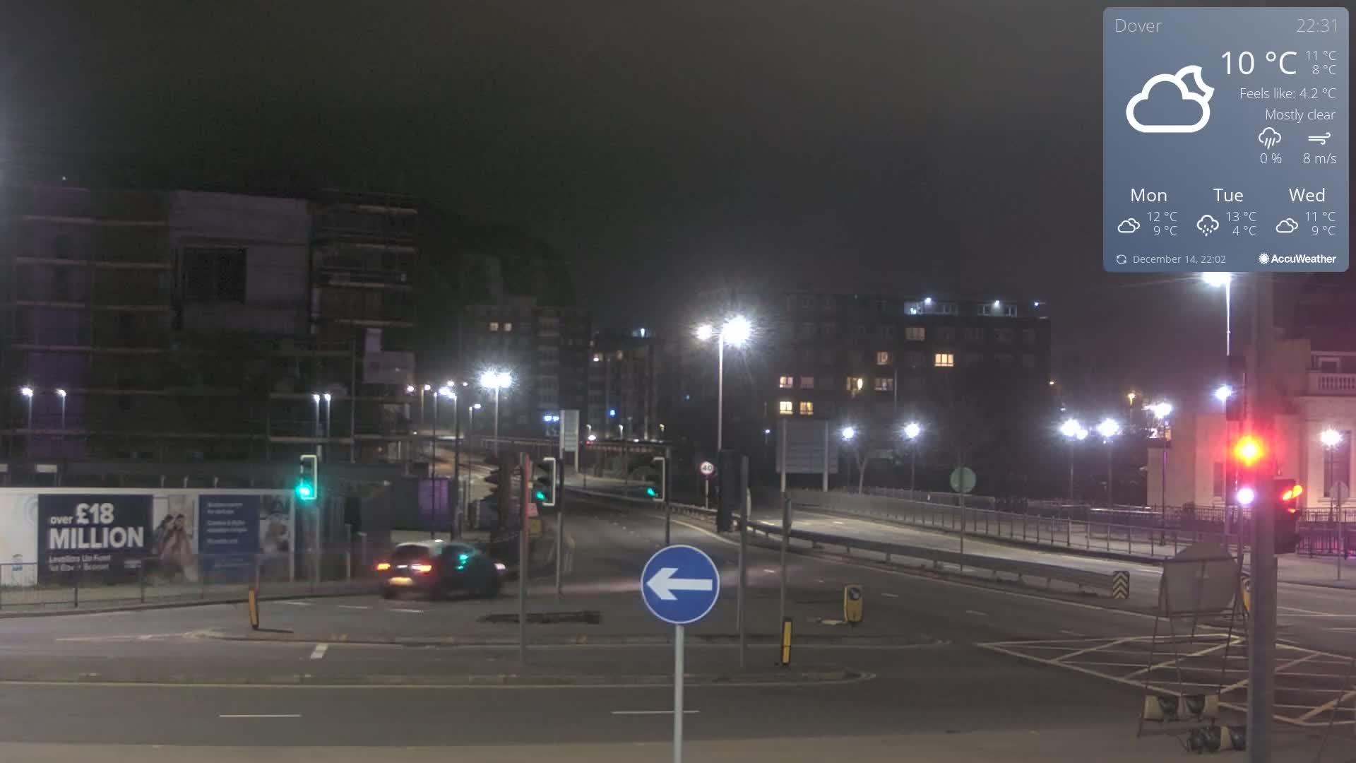 An urban street scene is heavily obscured by dense fog or mist, revealing only the faint outlines of distant buildings and the diffused lights of vehicles on the road.