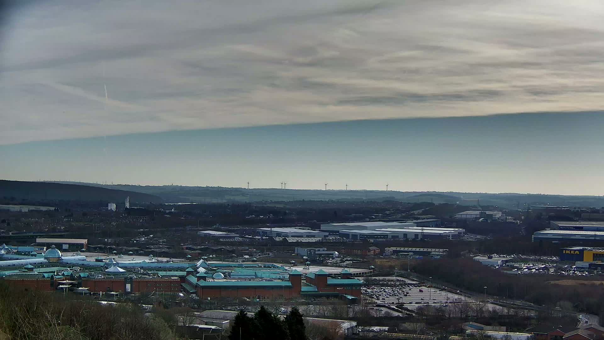 A sprawling cityscape with a large shopping mall in the foreground, industrial buildings in the middle ground, and a distant horizon featuring wind turbines under a mostly cloudy sky.
