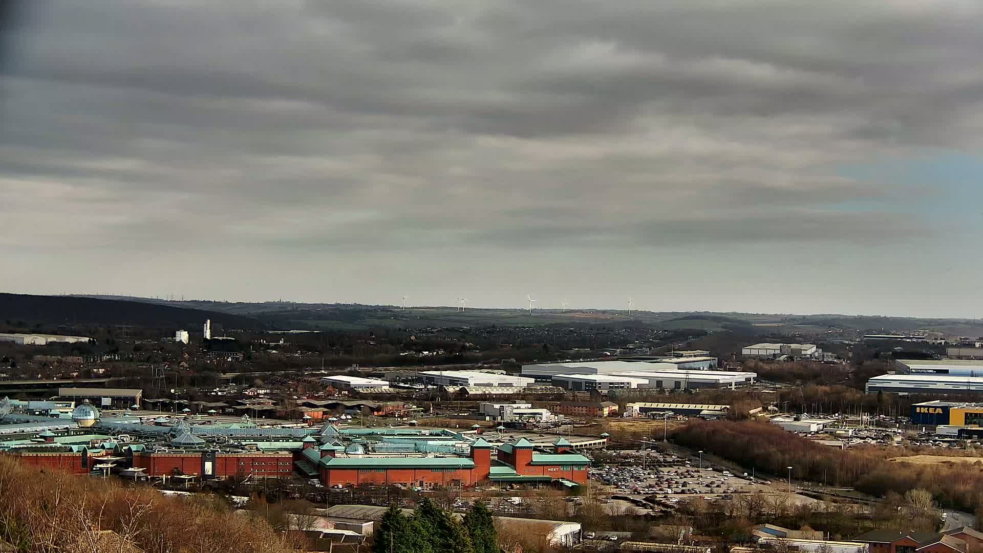 A sprawling shopping center and industrial complex are visible beneath a mostly cloudy sky, with wind turbines in the distance.