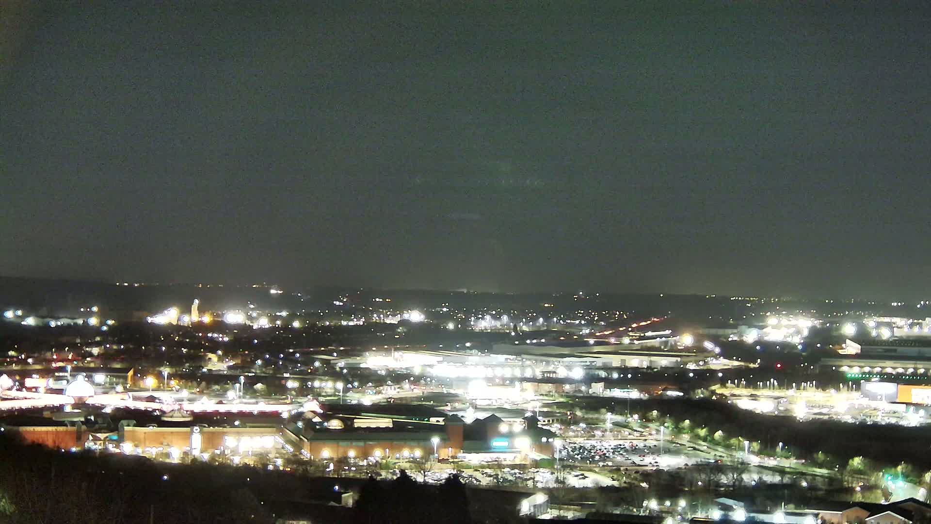 A nighttime aerial view of a sprawling city with illuminated buildings and parking lots under a dark, overcast sky.