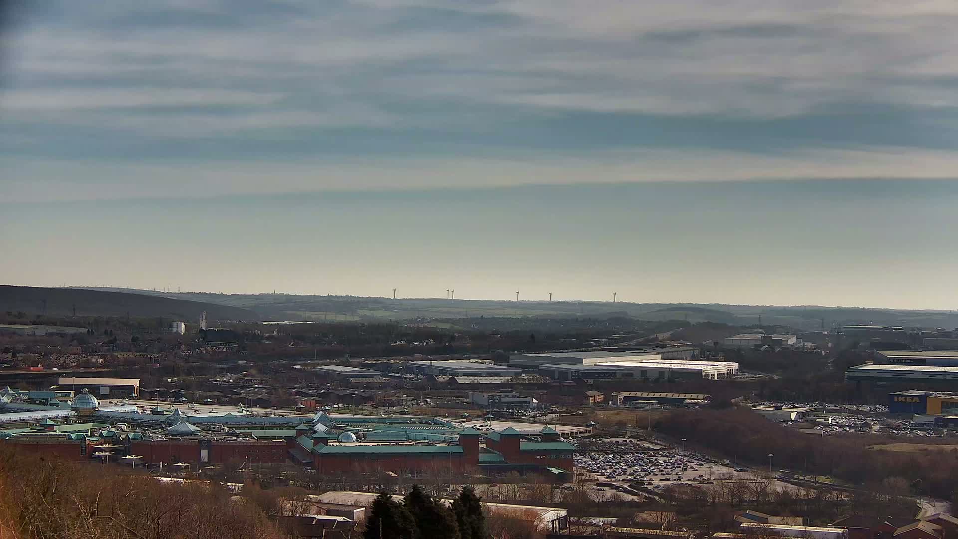 A sprawling shopping center and industrial area sits nestled in a valley under a mostly cloudy, bright sky, with distant wind turbines visible on the horizon.