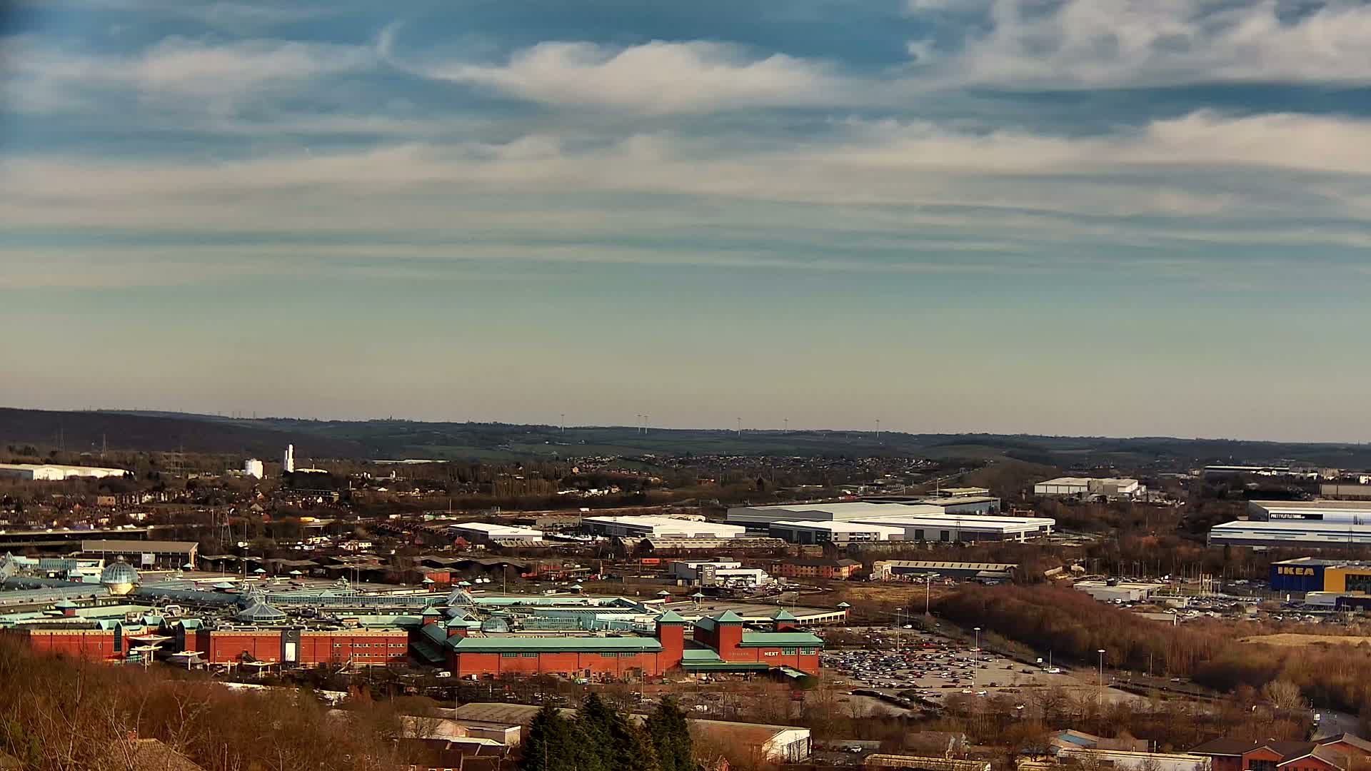 A sprawling shopping center with a large parking lot is visible from a high vantage point on a partly cloudy day, surrounded by industrial buildings and residential areas stretching to a distant horizon.