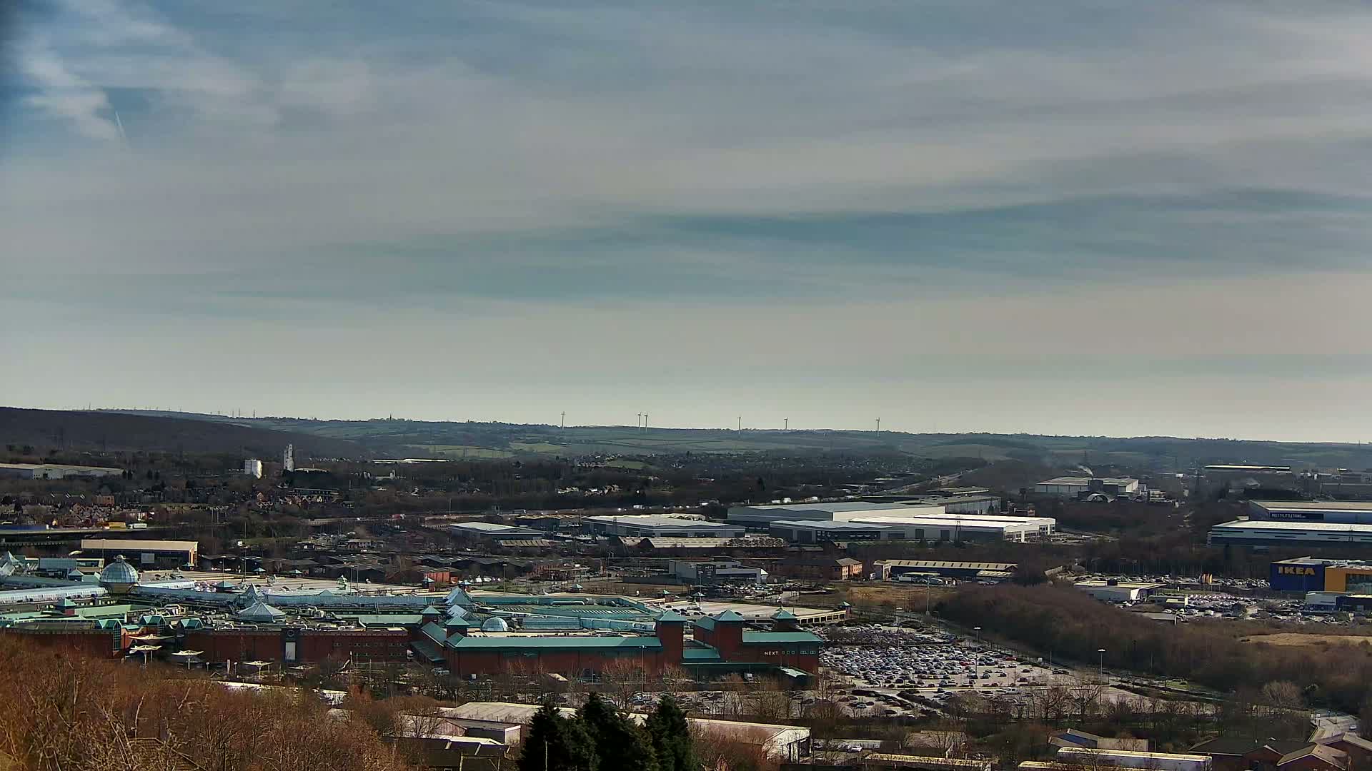 A large shopping mall with a teal roof is situated amongst industrial buildings and parking lots under a mostly cloudy sky.
