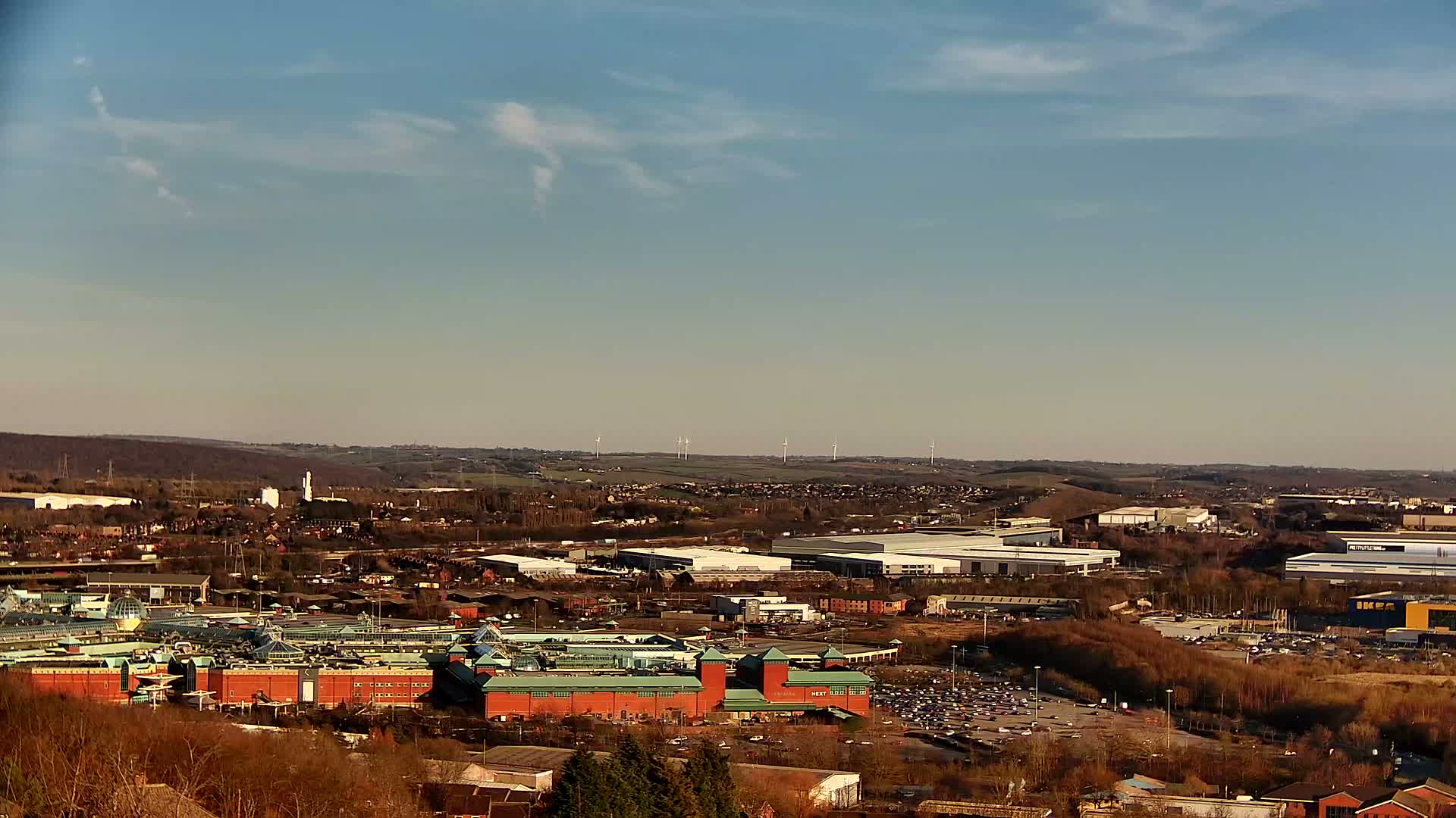 A large shopping mall with reddish-brown buildings and a green roof is situated in a sprawling landscape under a mostly clear, sunny sky with a few wispy clouds.