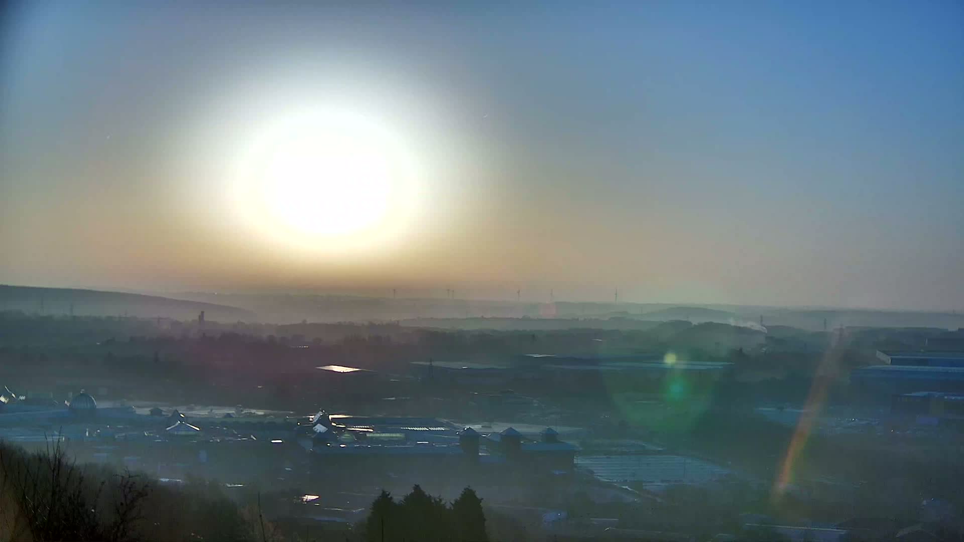 A hazy sunrise casts a bright glow over a distant industrial landscape, with a layer of fog or mist obscuring details.