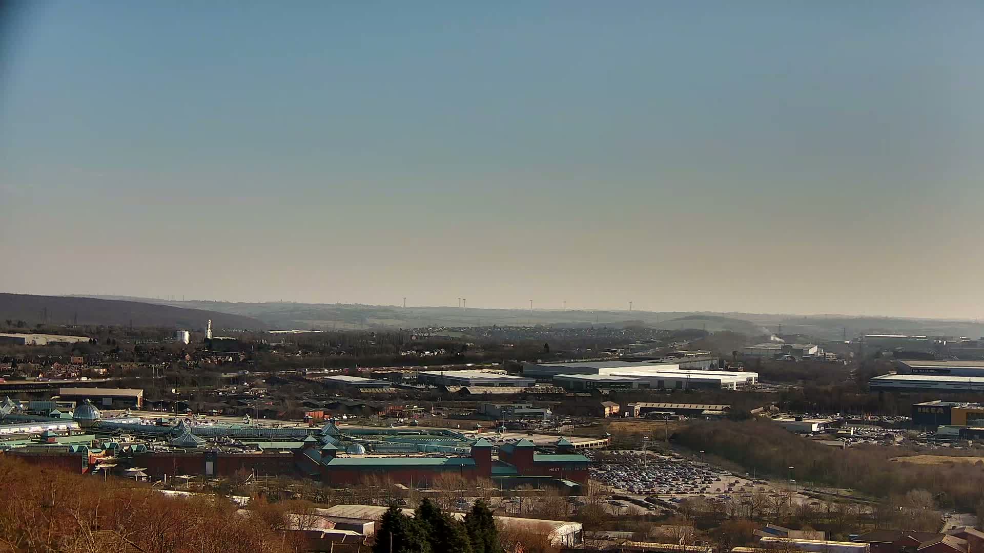 A sprawling suburban landscape with a large shopping mall and industrial buildings under a clear, sunny sky.