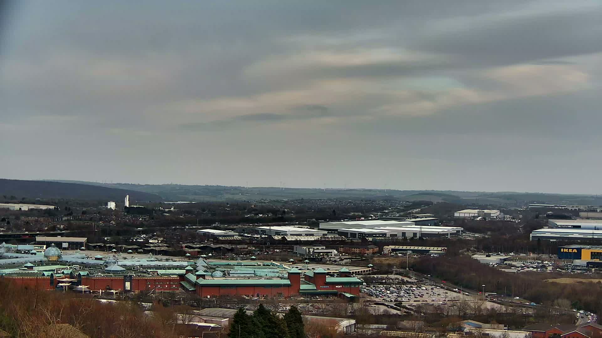 A sprawling cityscape under a cloudy sky features a large shopping mall and numerous industrial buildings, with a parking lot visible in the foreground.