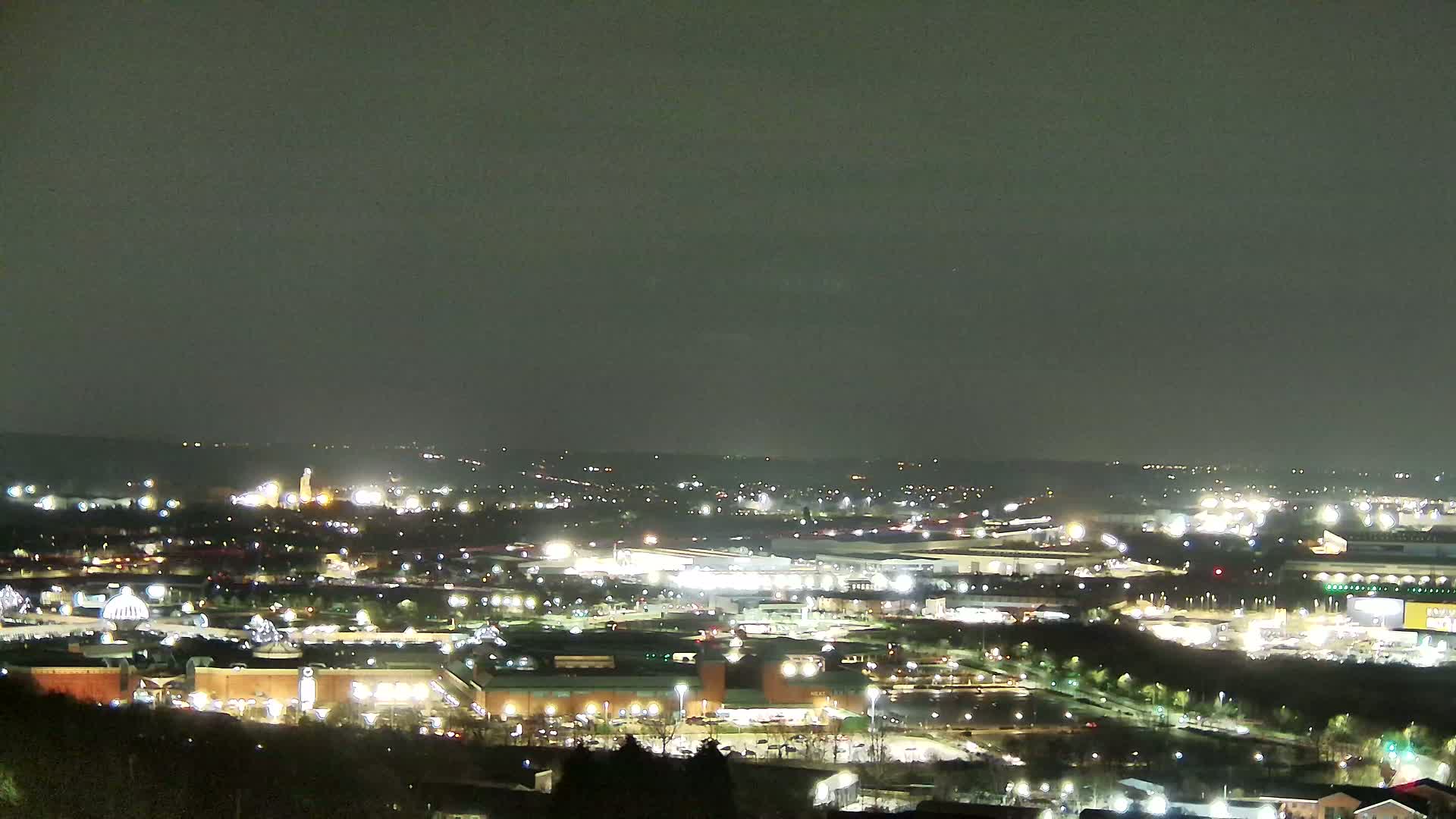 A nighttime long shot shows a sprawling cityscape with numerous illuminated buildings and roads under a dark, overcast sky.
