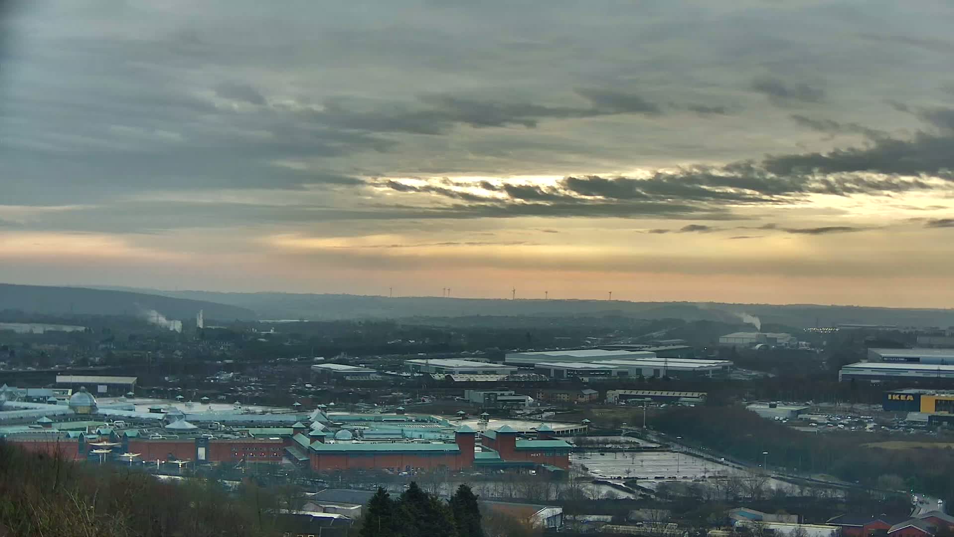 A sprawling industrial area and large shopping mall are visible from a high vantage point under a mostly cloudy sky with a hint of sunset.