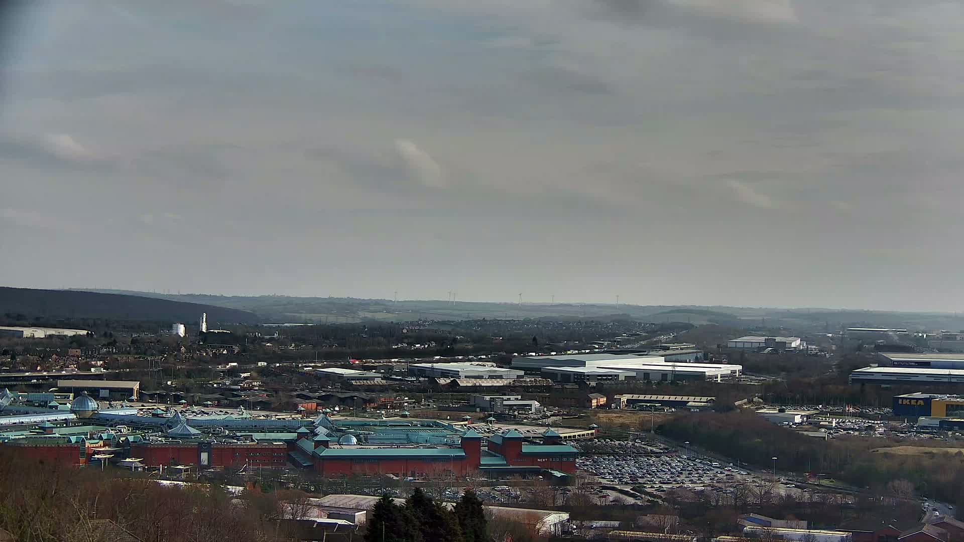 A large shopping mall with a teal roof is situated in a sprawling suburban area under a cloudy sky.