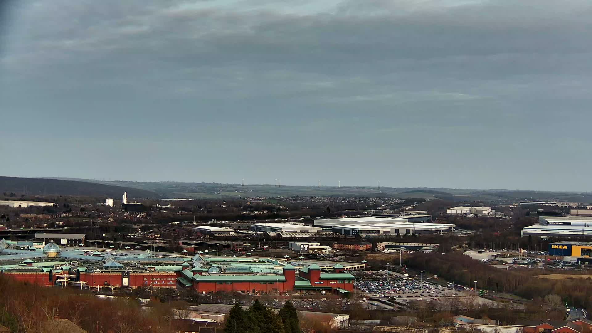 A large shopping mall and surrounding industrial buildings are seen from an elevated vantage point on an overcast day, with wind turbines visible in the distance.