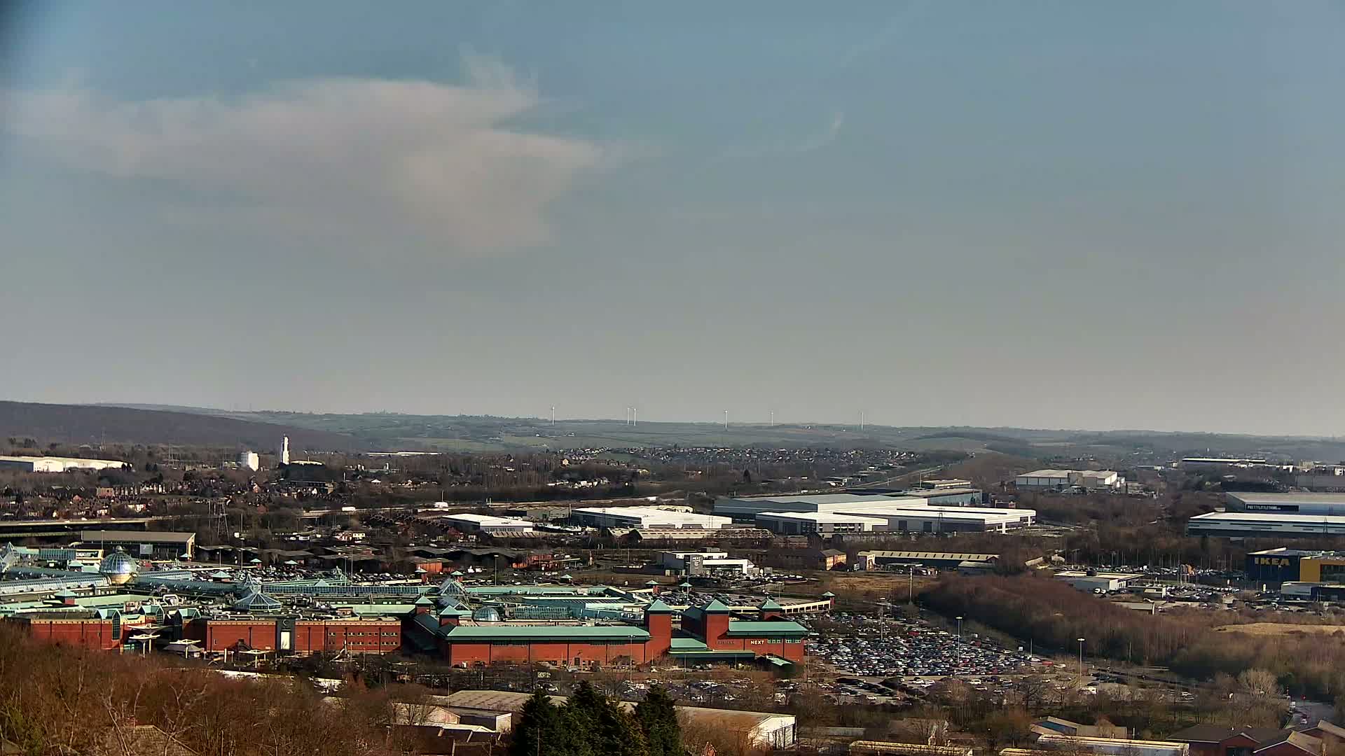 A large shopping mall with a sprawling parking lot is visible in the foreground, under a mostly clear sky with a few clouds, extending to a horizon filled with industrial buildings and low-lying hills in the distance.