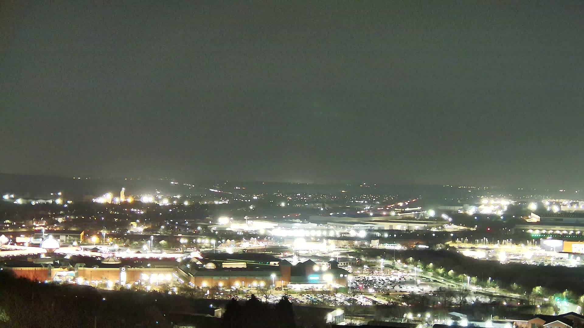 A nighttime, high-angle view of a sprawling shopping center and surrounding city lights under a dark, overcast sky.