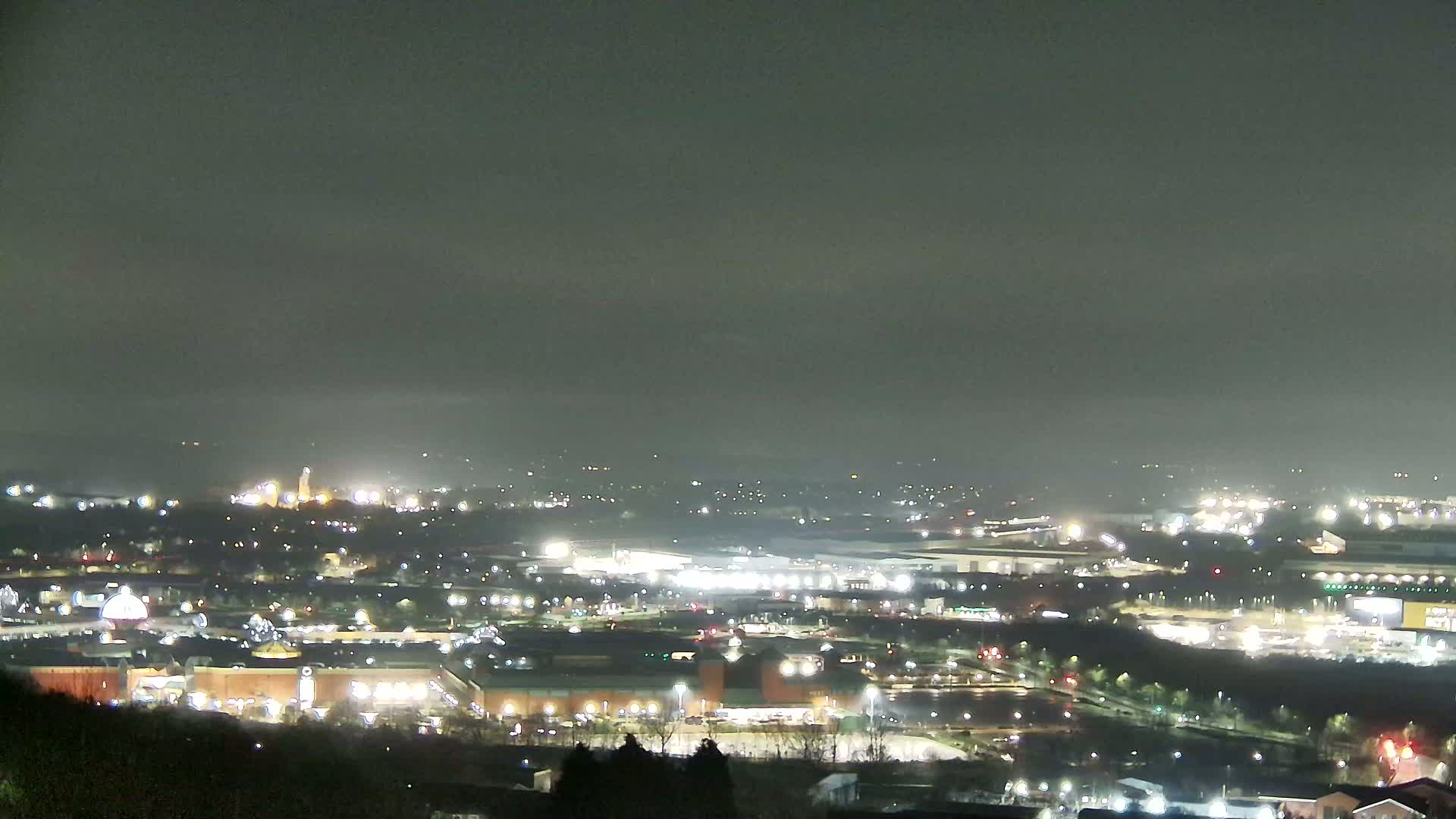 A nighttime view of a sprawling city landscape under a hazy sky, showing numerous illuminated buildings and roadways.
