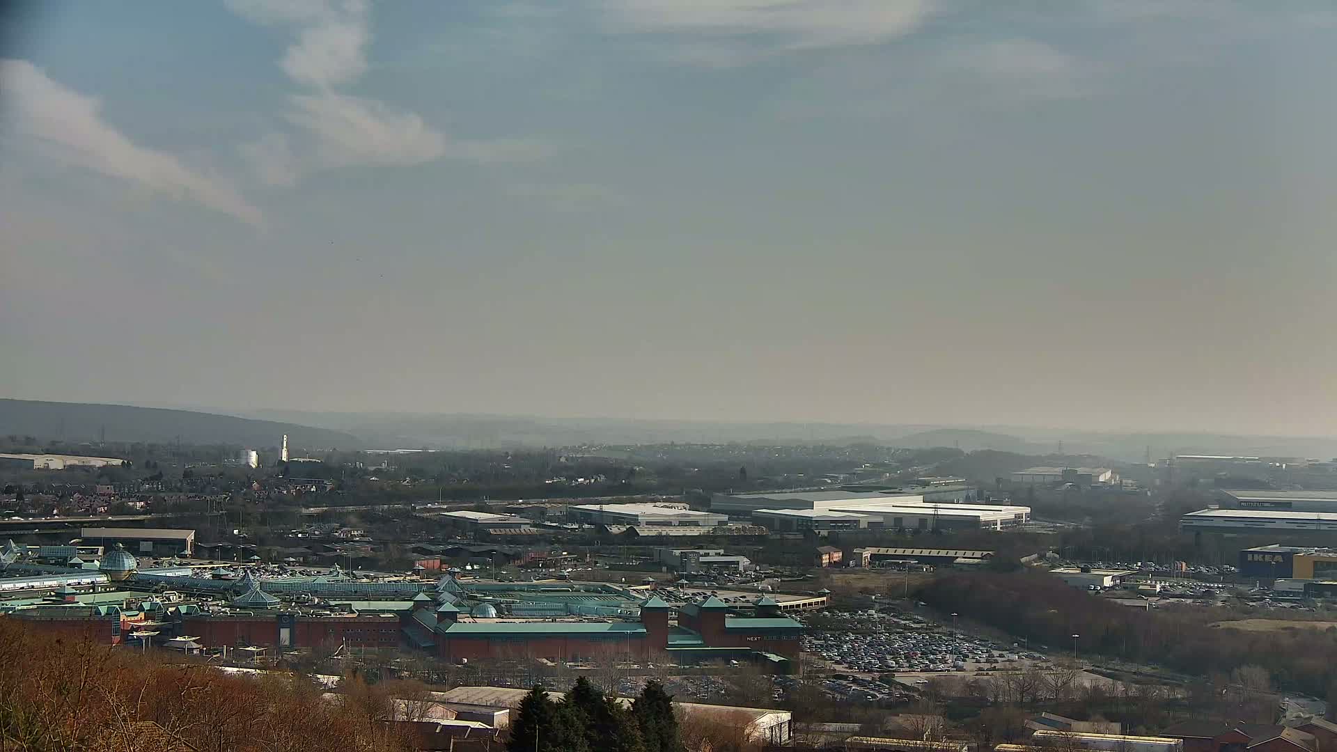 A hazy, sunny day reveals a sprawling cityscape with a large shopping mall at its center, surrounded by industrial buildings and parking lots, viewed from a high vantage point.