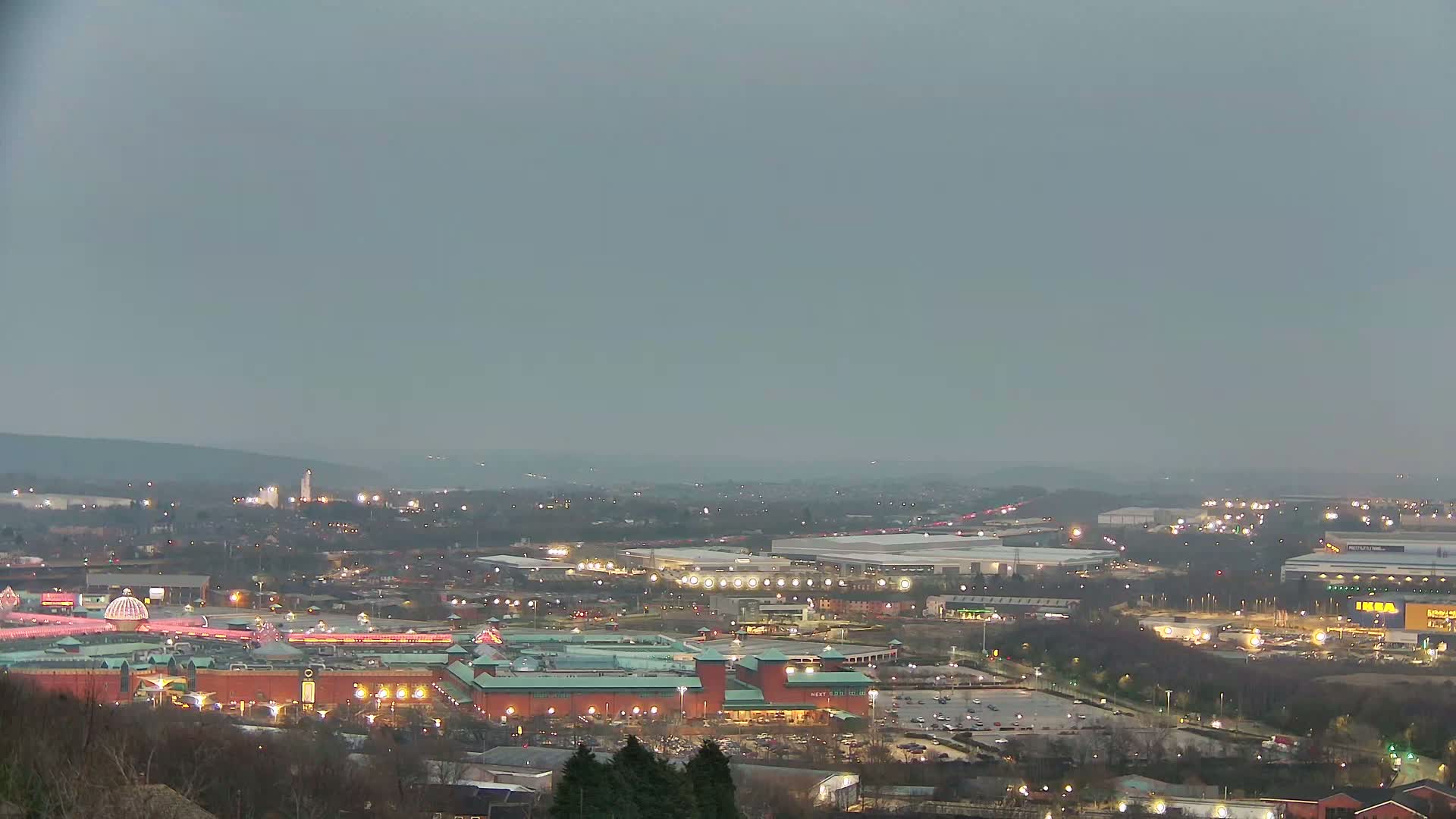 A sprawling cityscape at dusk or dawn under a hazy, overcast sky, featuring a large shopping mall and surrounding industrial areas illuminated with lights.