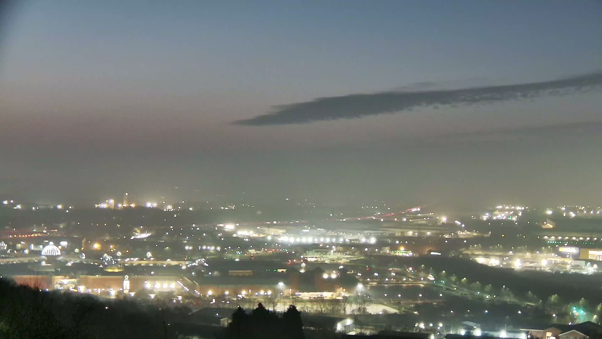 A hazy night view shows a sprawling city landscape illuminated by numerous lights, with a long, dark cloud partially obscuring the sky.
