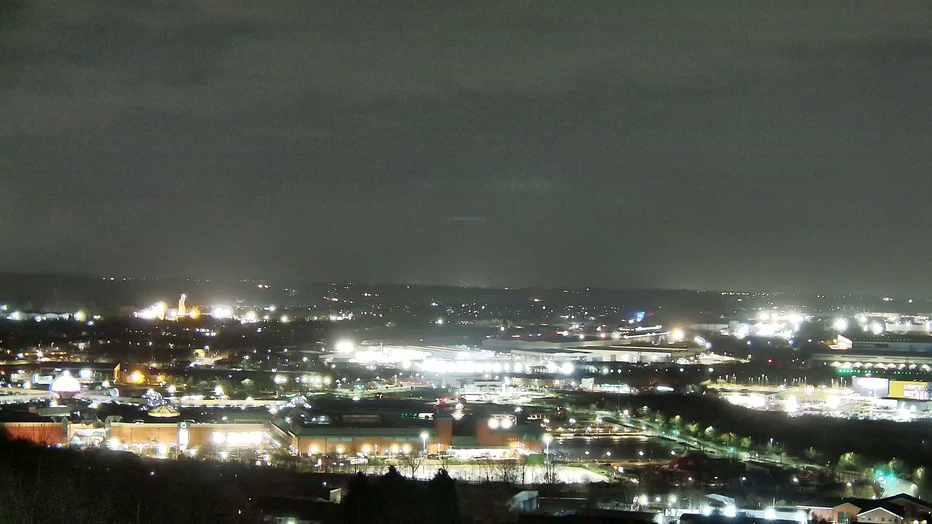 A nighttime, overcast view shows a sprawling complex of brightly lit buildings and parking lots, surrounded by dark trees and a distant, dimly lit cityscape under a dark sky.