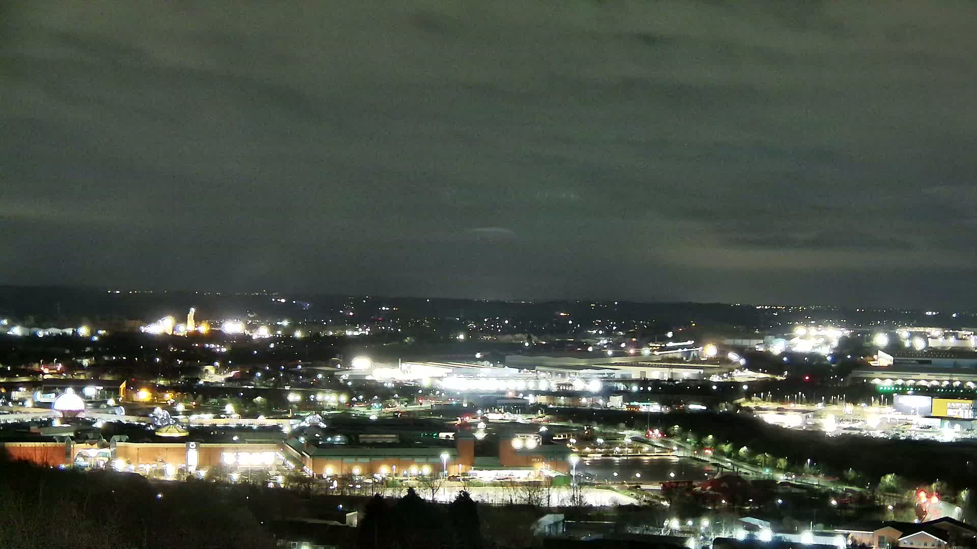 A nighttime view of a sprawling city illuminated by numerous lights under a mostly cloudy sky.