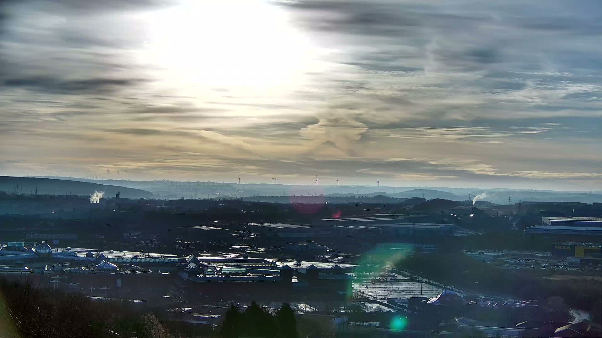 A hazy, sunlit day reveals a sprawling industrial landscape punctuated by a large shopping center, wind turbines in the distance, and wisps of smoke rising from factories.