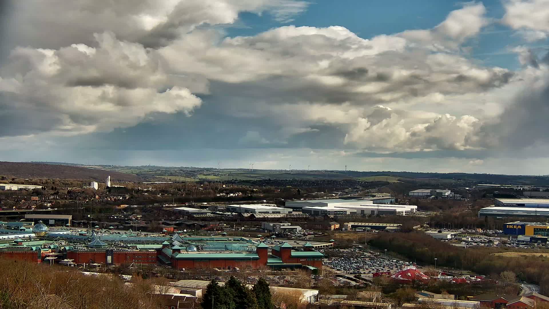 A large shopping mall with a glass roof is situated in a sprawling suburban landscape under a partly cloudy sky.