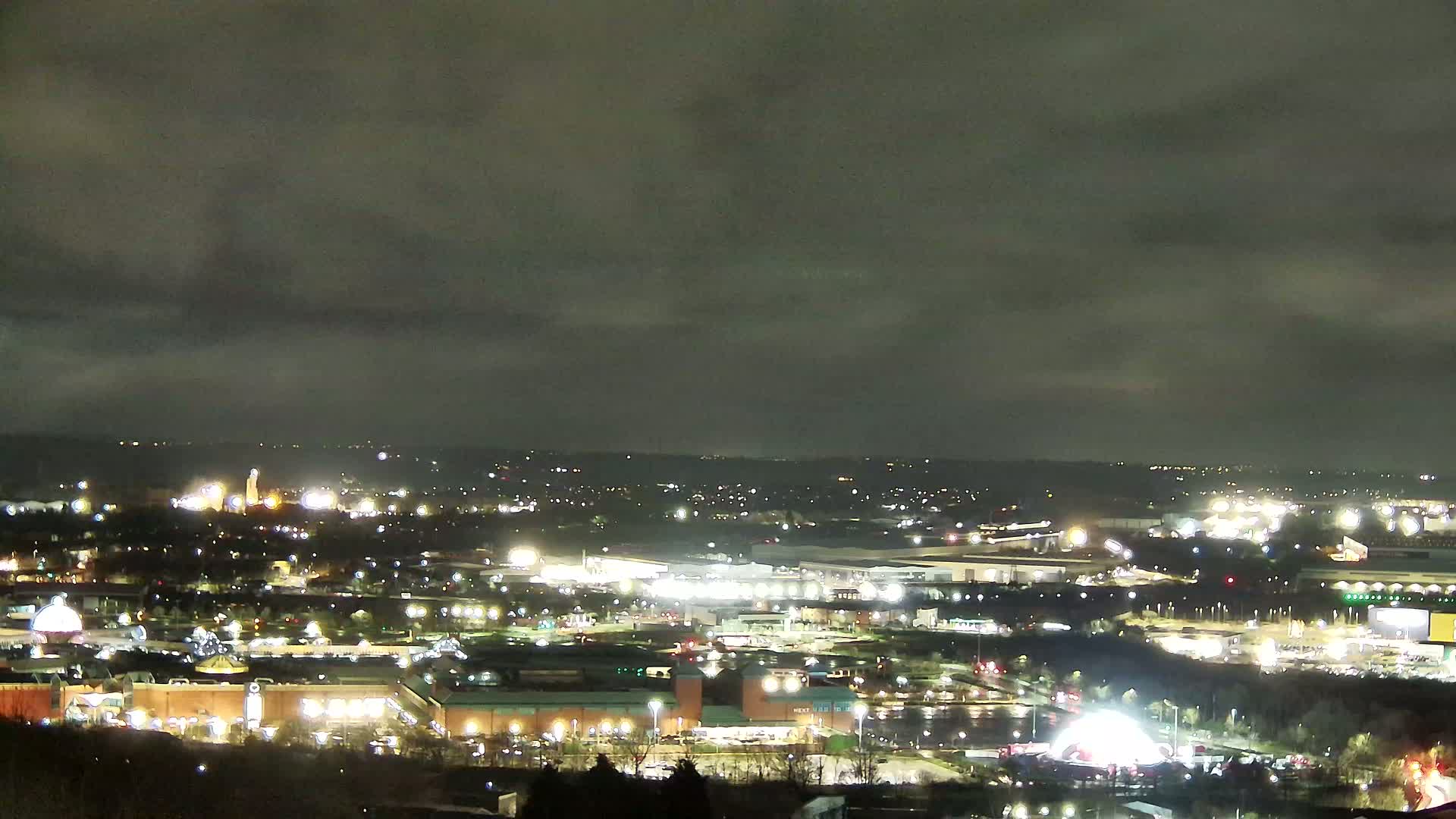A nighttime, overcast view of a sprawling city landscape illuminated by numerous lights, showing a mix of residential, commercial, and industrial areas.