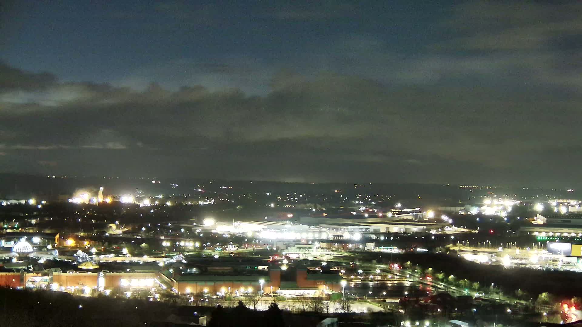 A nighttime, high-angle view of a city illuminated with numerous lights under a partly cloudy sky.