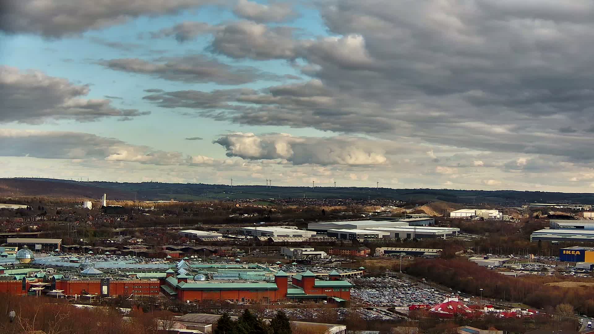 A large shopping center with expansive parking lots is situated amongst industrial buildings and residential areas under a partly cloudy sky.