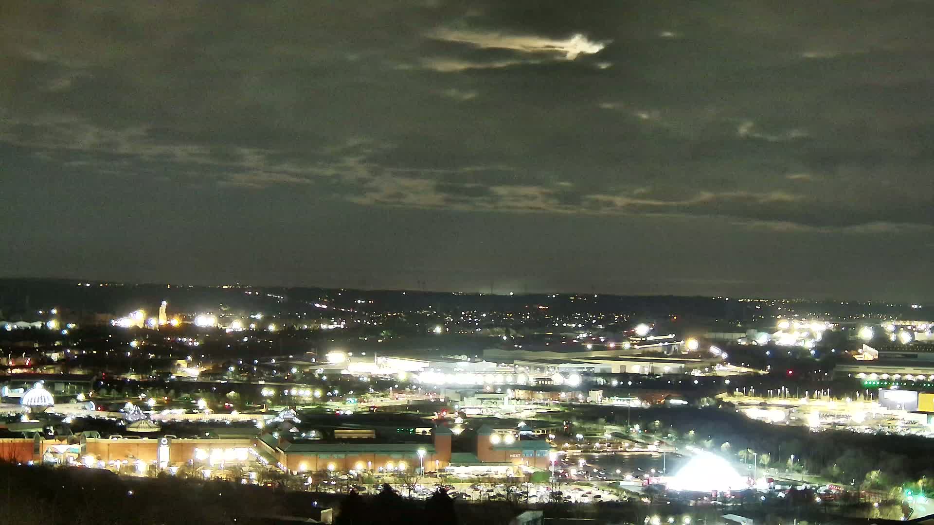A nighttime, partly cloudy view of a sprawling city illuminated by numerous lights, featuring large retail spaces and parking lots.