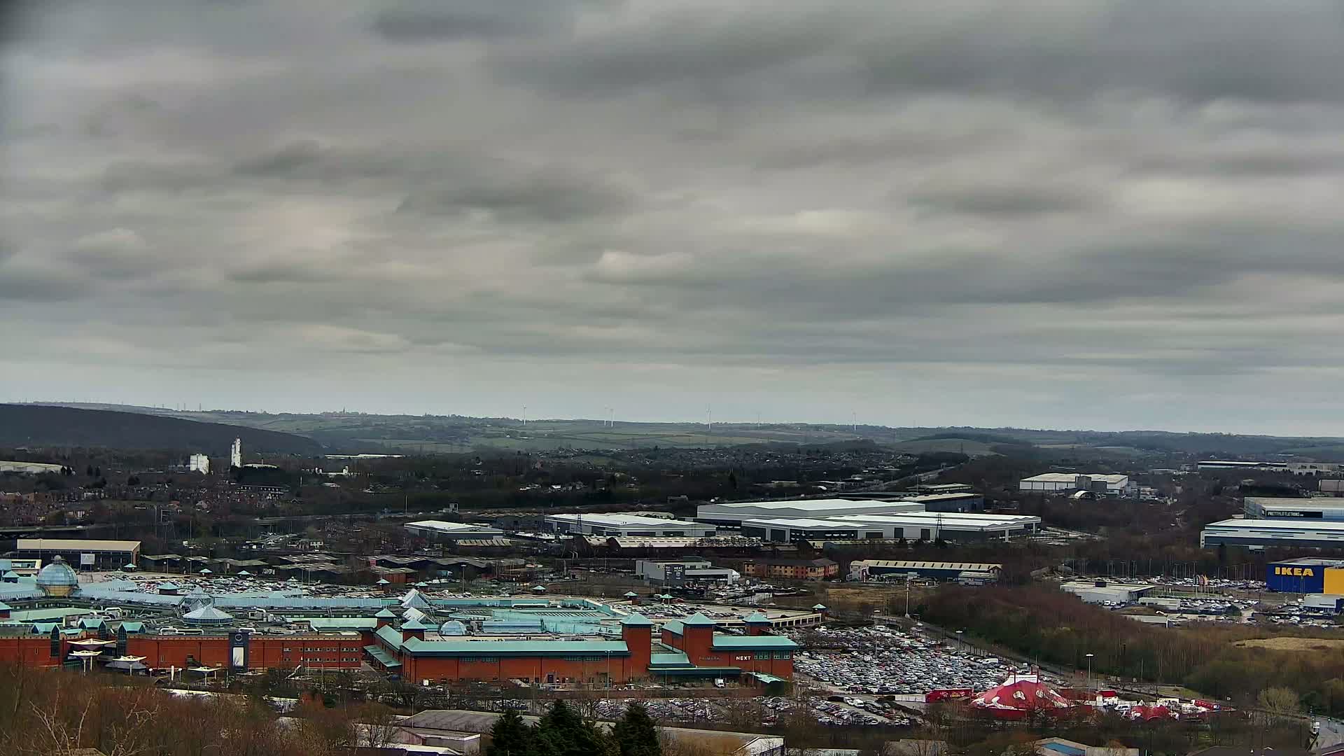 A large shopping center and surrounding industrial areas are seen from a distance under a cloudy sky.