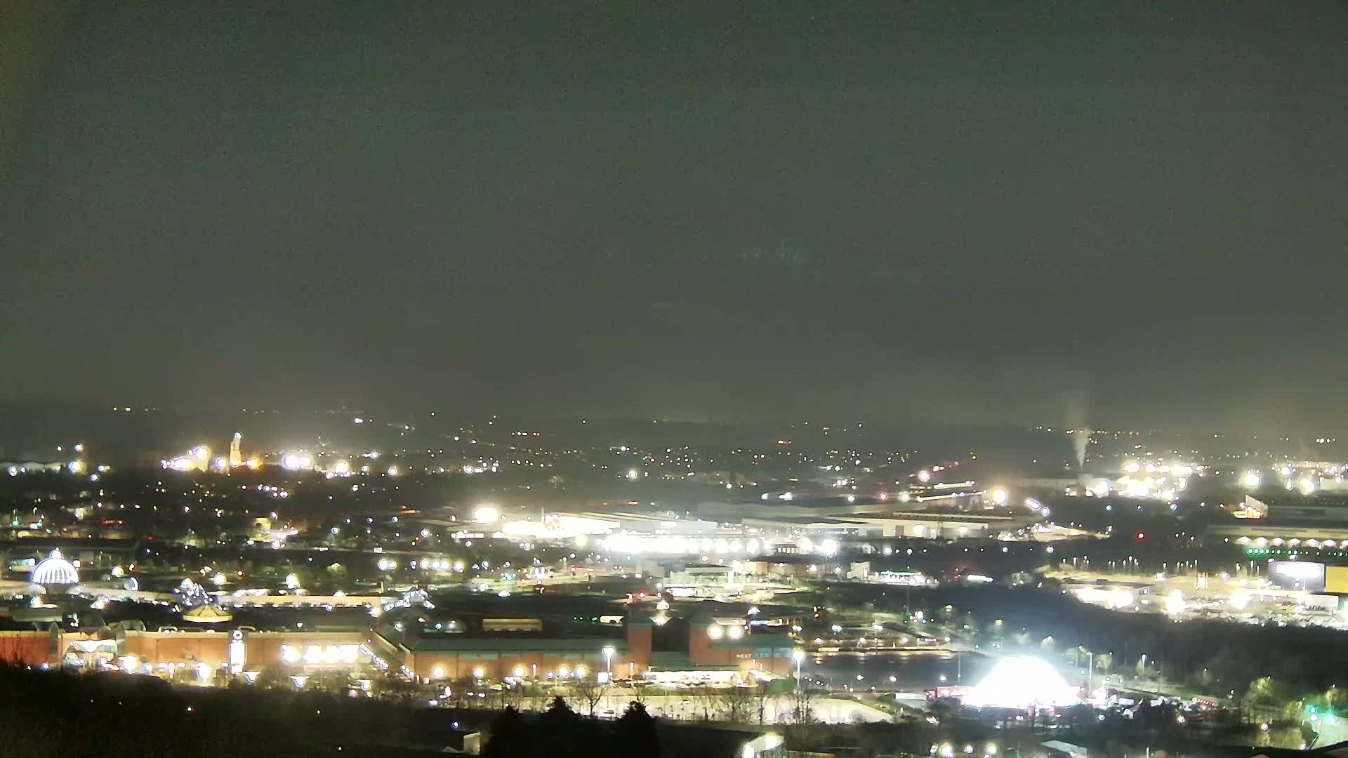 A nighttime, long-range view of a sprawling city and its illuminated buildings, under a hazy sky.