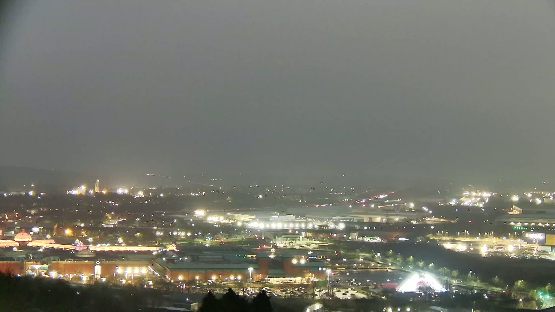 A nighttime, hazy view of a large shopping center and surrounding city lights, seen from an elevated vantage point.