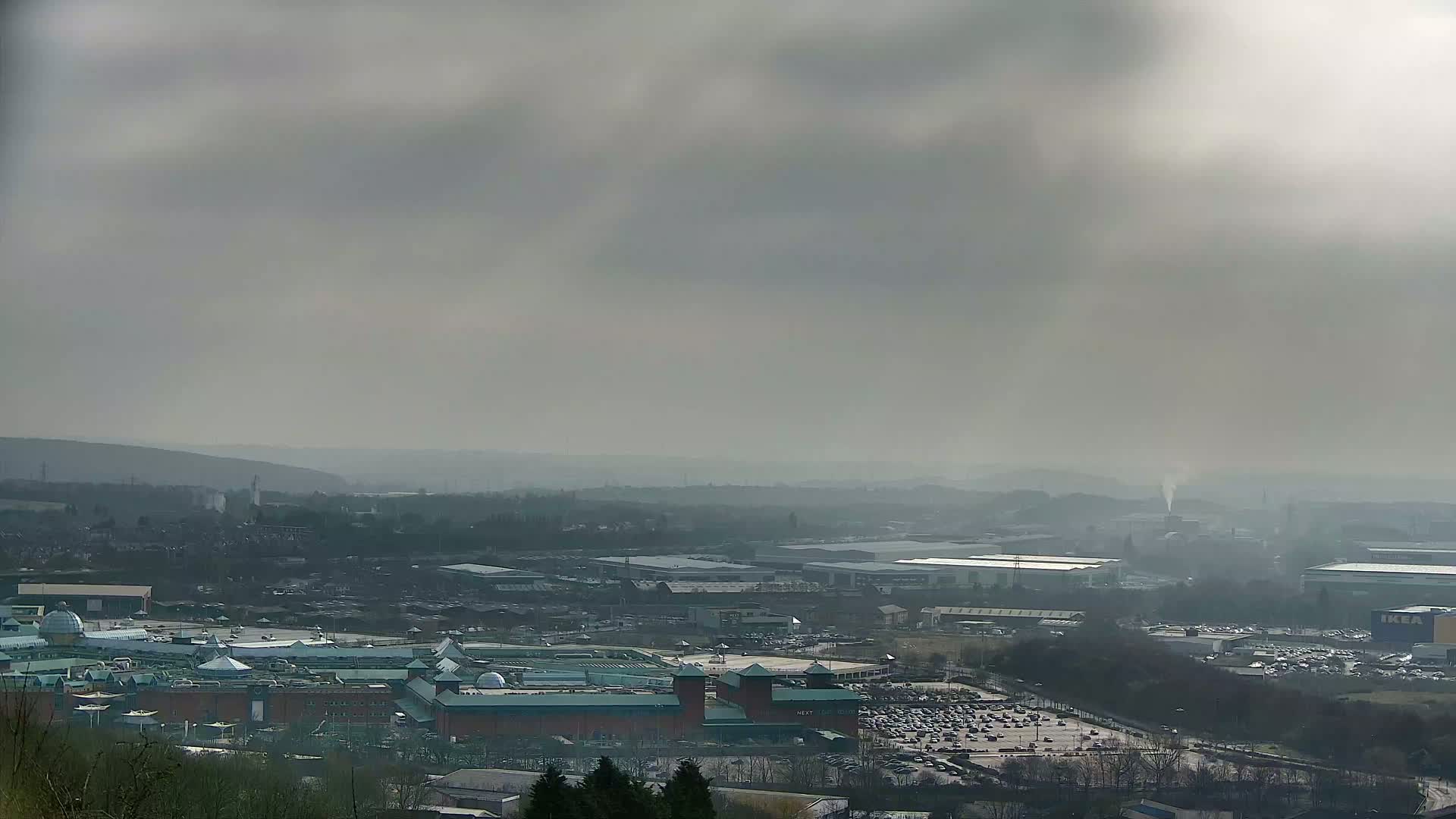 A hazy, overcast day reveals a sprawling cityscape with a large shopping mall at its center, surrounded by industrial areas and parking lots.