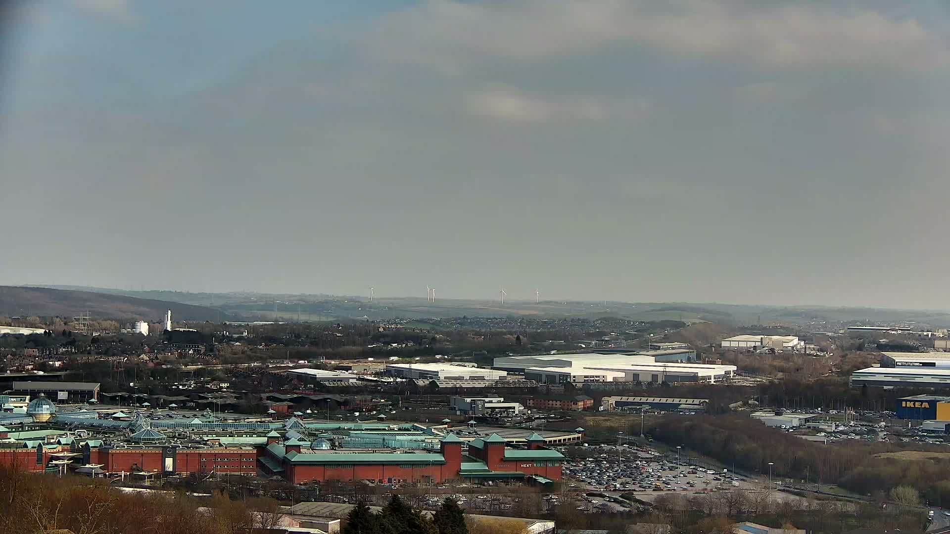 A sprawling cityscape under a hazy sky, featuring a large shopping mall and industrial areas, with wind turbines visible in the distance.