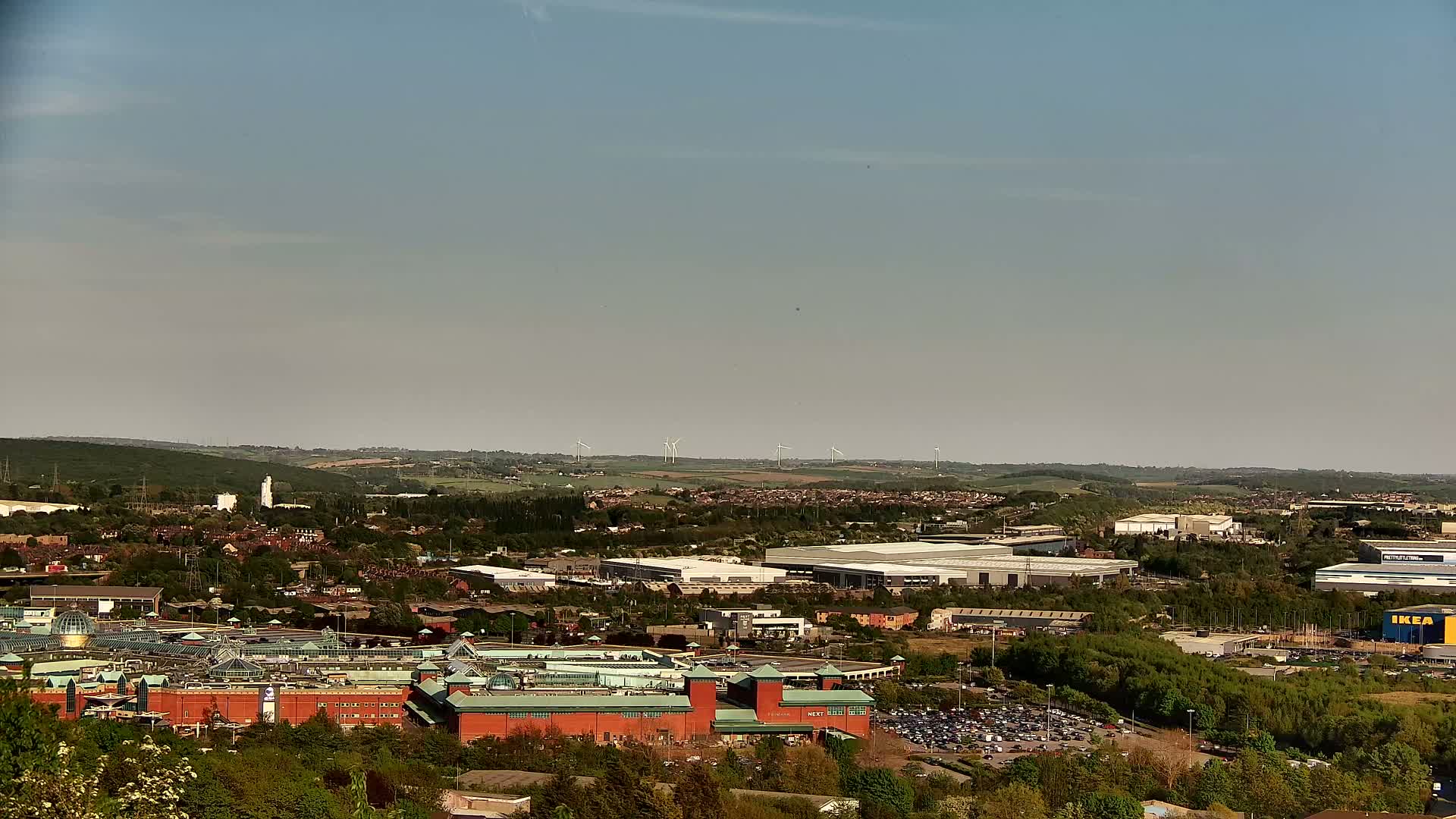 A sprawling suburban landscape under a clear, light-blue sky features a large shopping center, industrial buildings, and a line of wind turbines in the distance.