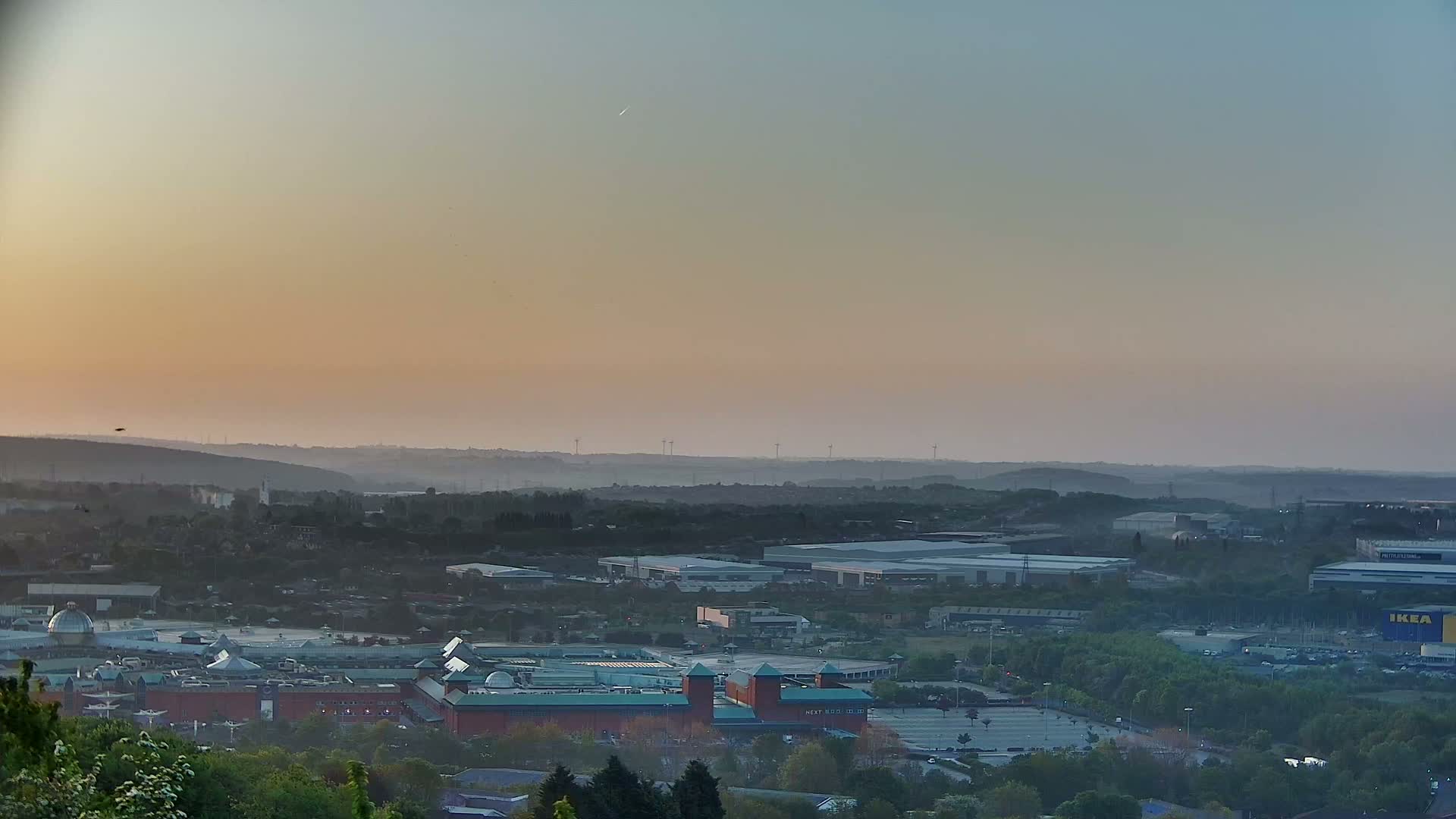 A hazy sunset illuminates a sprawling suburban landscape dotted with industrial buildings and a large shopping center.