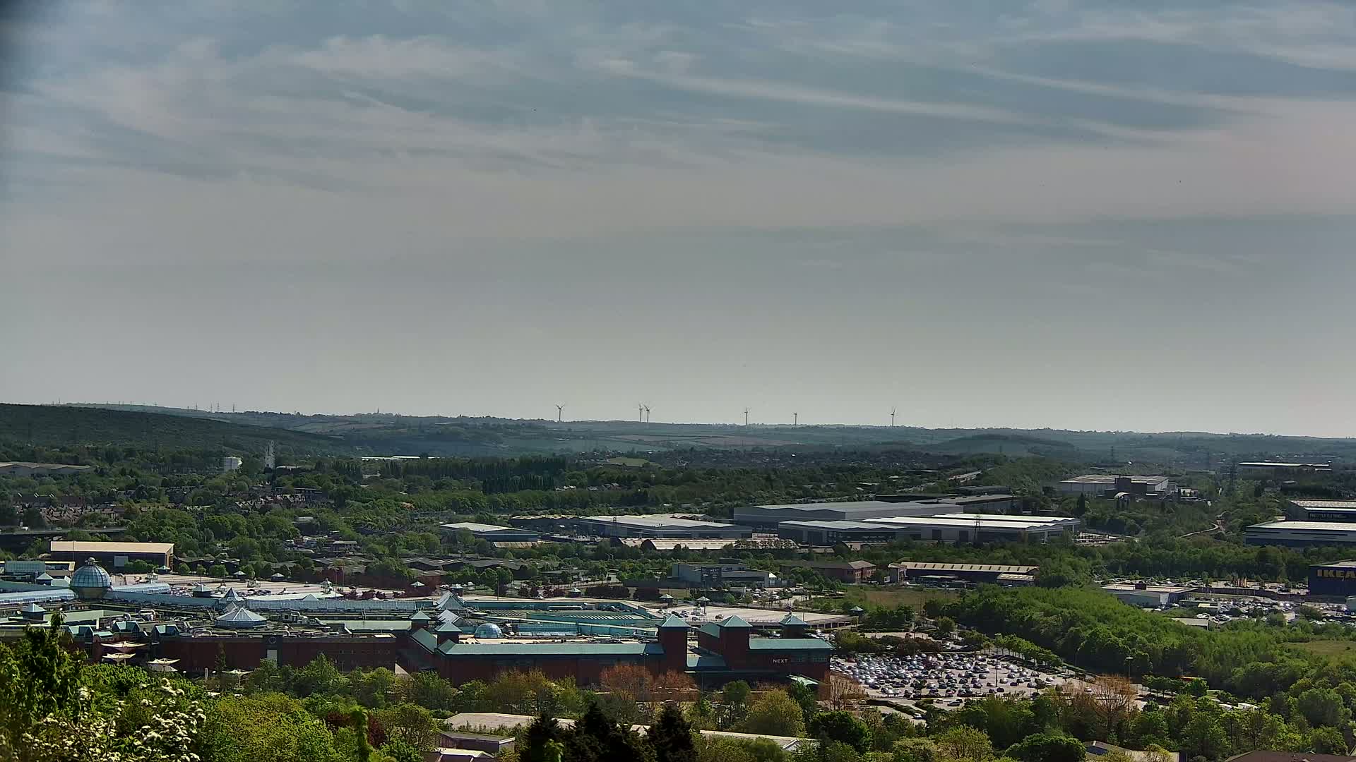 A sprawling shopping center with a large parking lot is nestled in a green valley under a mostly cloudy, but bright sky, with wind turbines visible in the distance.