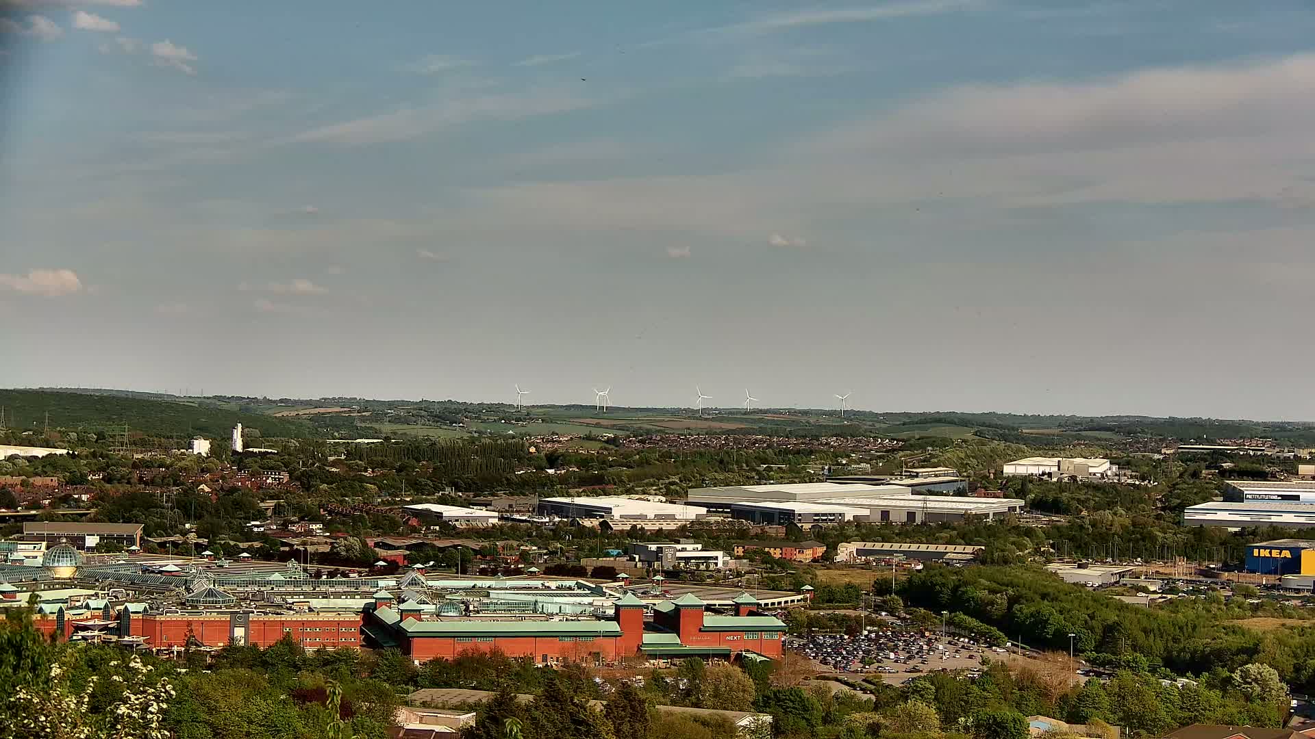 A sprawling suburban landscape under a partly cloudy sky features a large shopping center, industrial buildings, and a line of wind turbines in the distance.