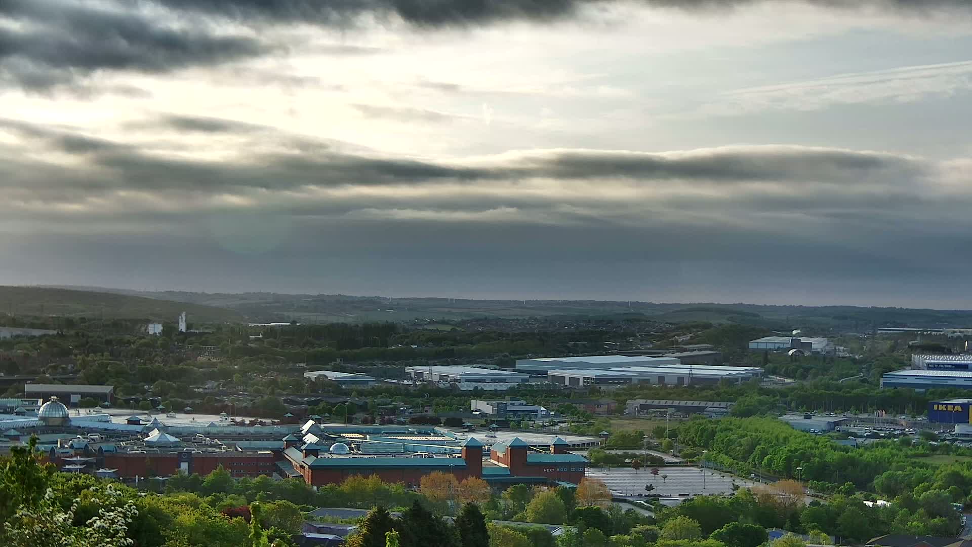 A sprawling shopping mall and industrial park are situated amidst a landscape of green trees and rolling hills under a mostly cloudy sky.