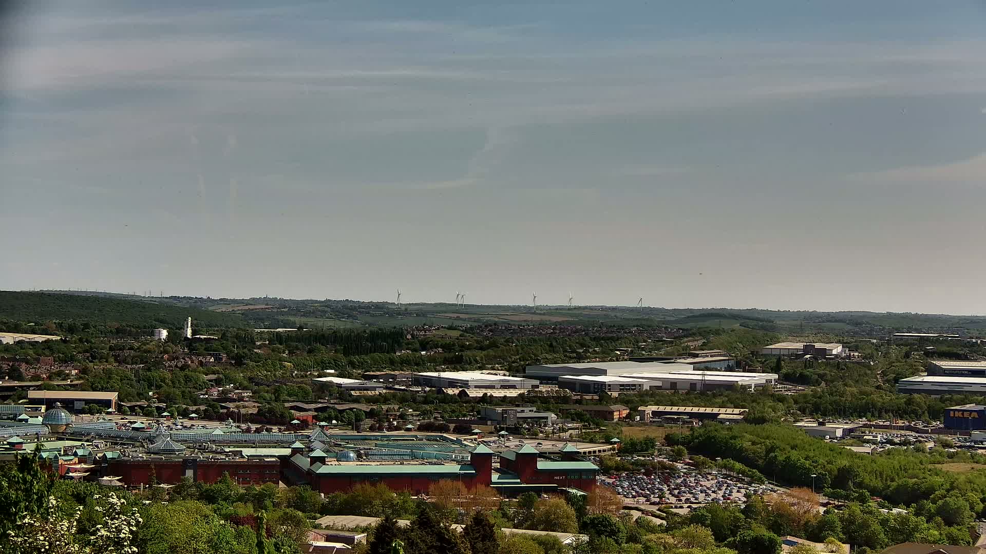 A sprawling shopping center with a red roof and glass atriums sits nestled among green trees and industrial buildings under a mostly clear, light-blue sky, with wind turbines visible in the distance.