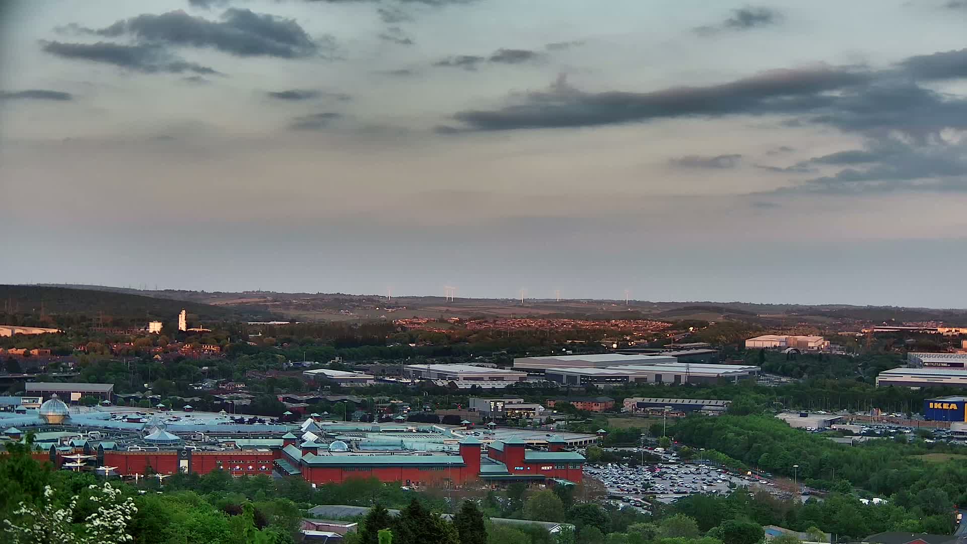 A large shopping mall and surrounding industrial buildings are visible from an elevated vantage point under a cloudy, dusky sky.