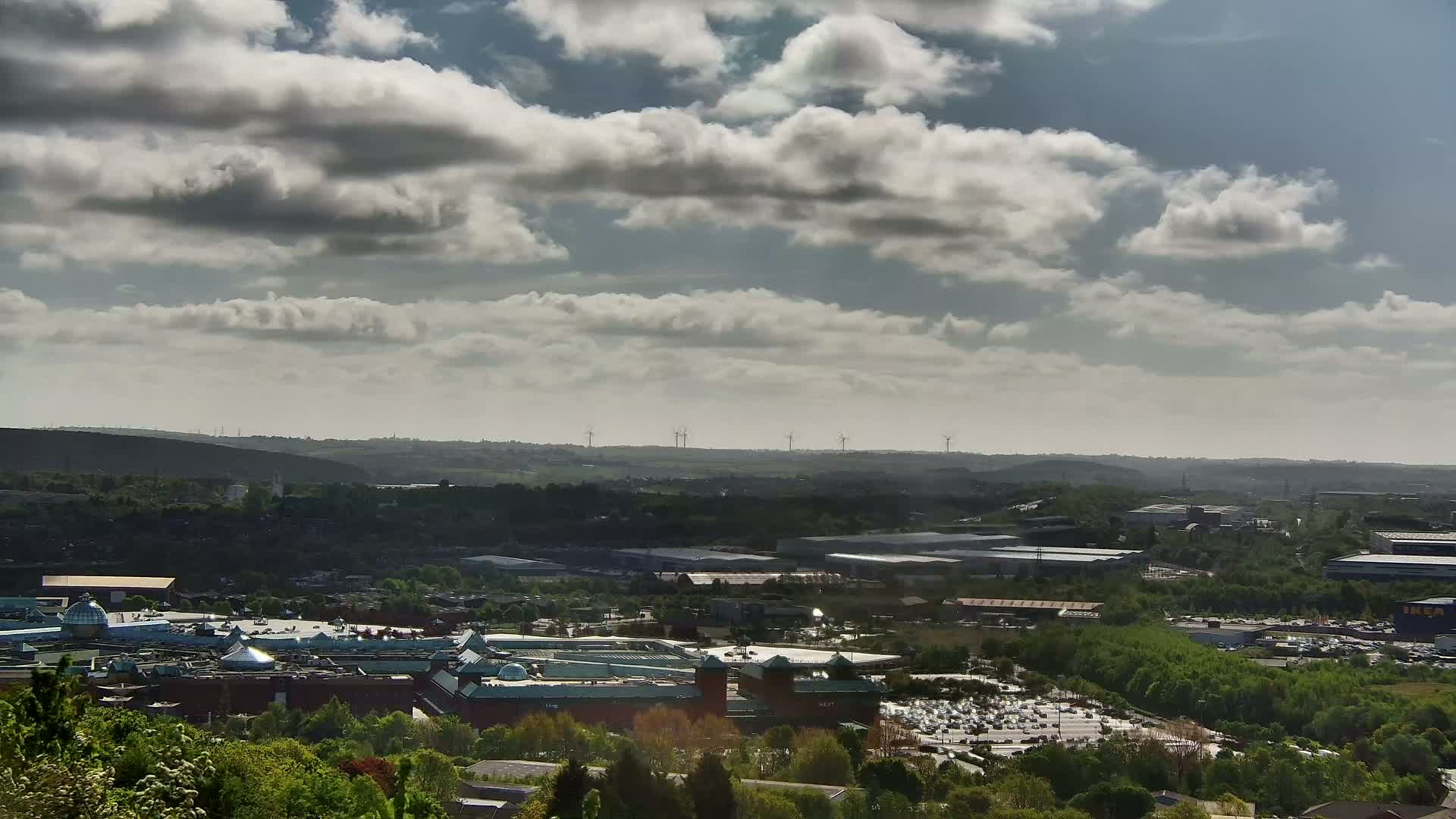 A sprawling shopping center and industrial park are nestled in a verdant valley under a partly cloudy sky, with wind turbines visible in the distance.