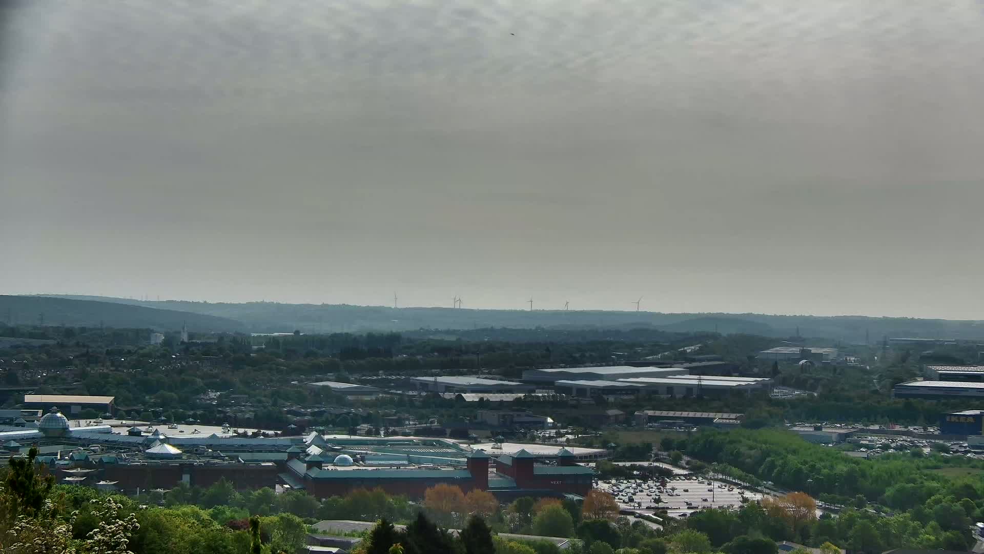 A sprawling shopping center and industrial park are visible beneath a hazy, overcast sky, with wind turbines in the distance.