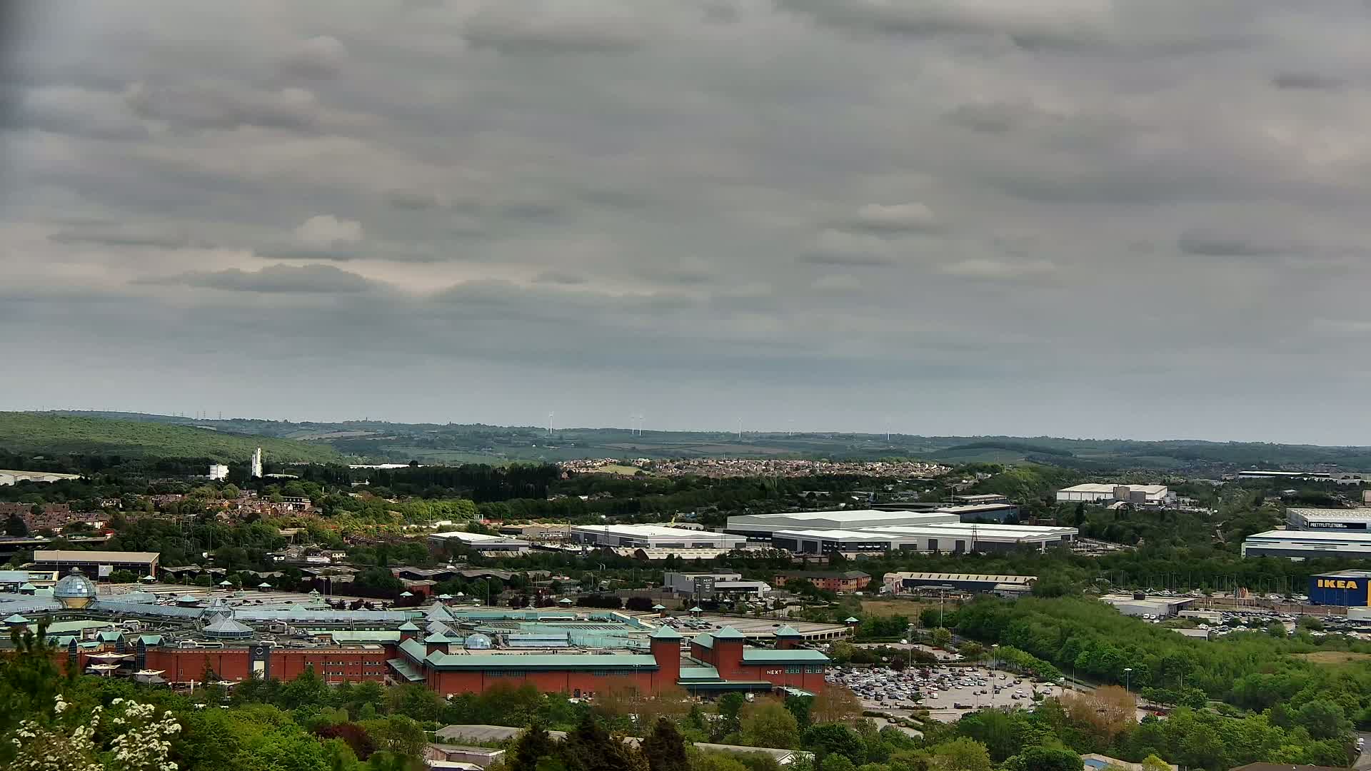 A large shopping center and surrounding industrial buildings are visible from an elevated vantage point under a cloudy sky.
