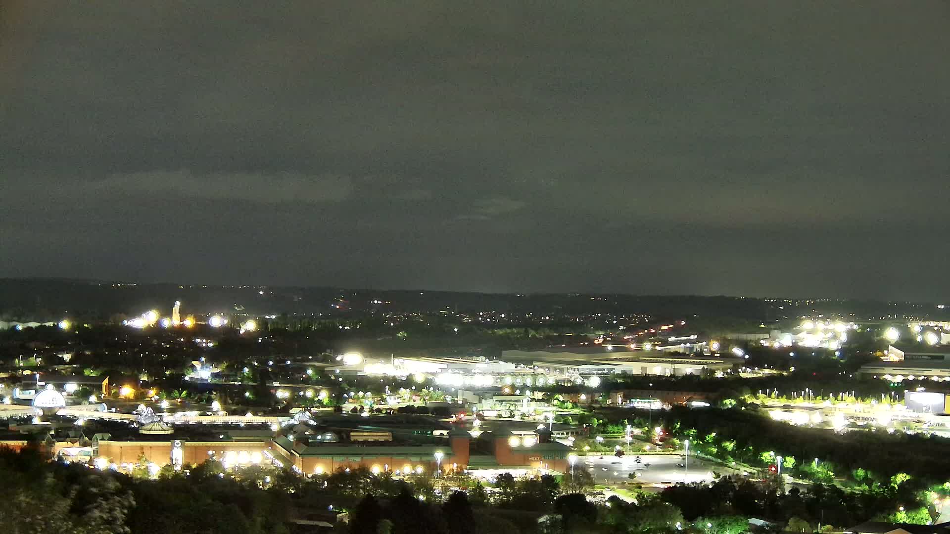 A nighttime, overcast view of a sprawling commercial area and surrounding landscape, illuminated by artificial lights.