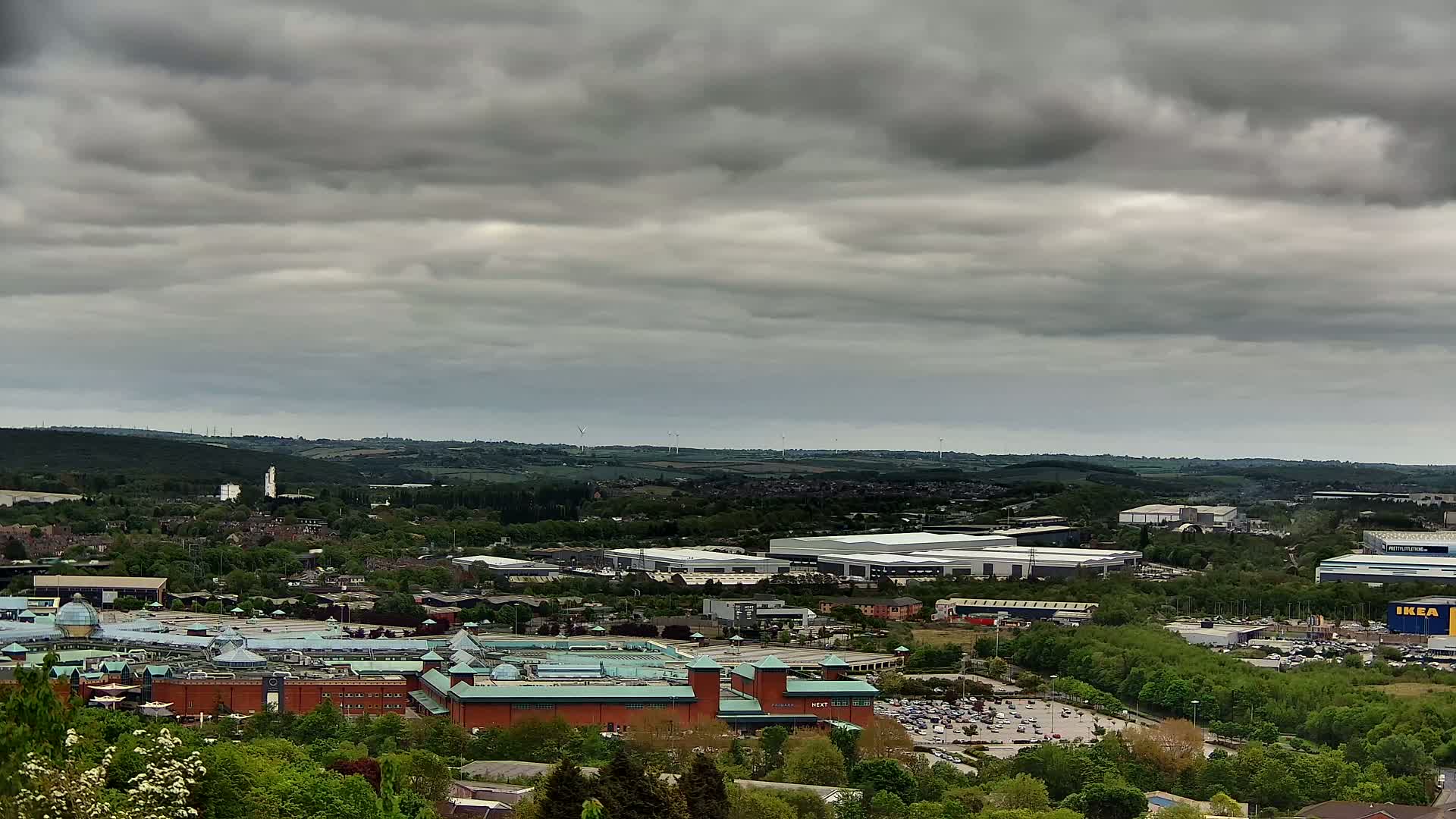 A sprawling shopping center and industrial park are visible beneath an overcast sky, nestled within a verdant landscape dotted with trees and buildings.