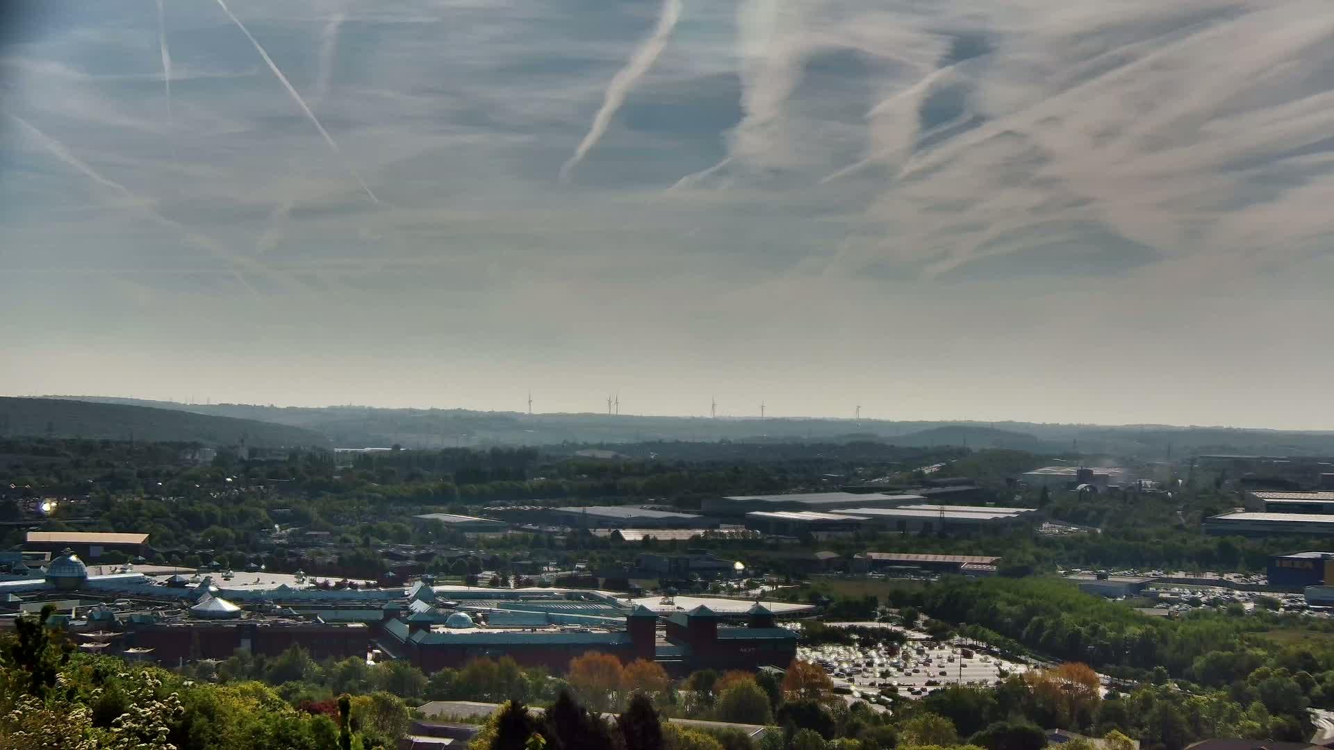 A sprawling shopping center sits nestled in a green valley, under a hazy sky streaked with contrails.