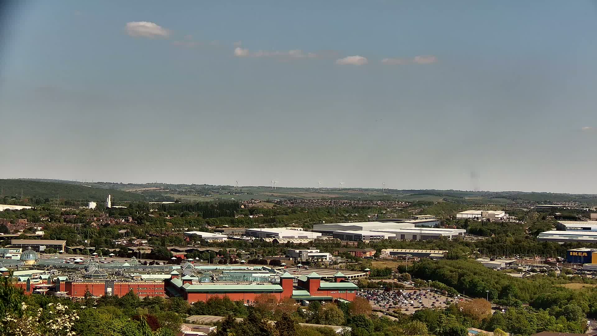 A sprawling shopping center and industrial park are visible from an elevated vantage point on a partly sunny day, with a line of wind turbines in the distance.