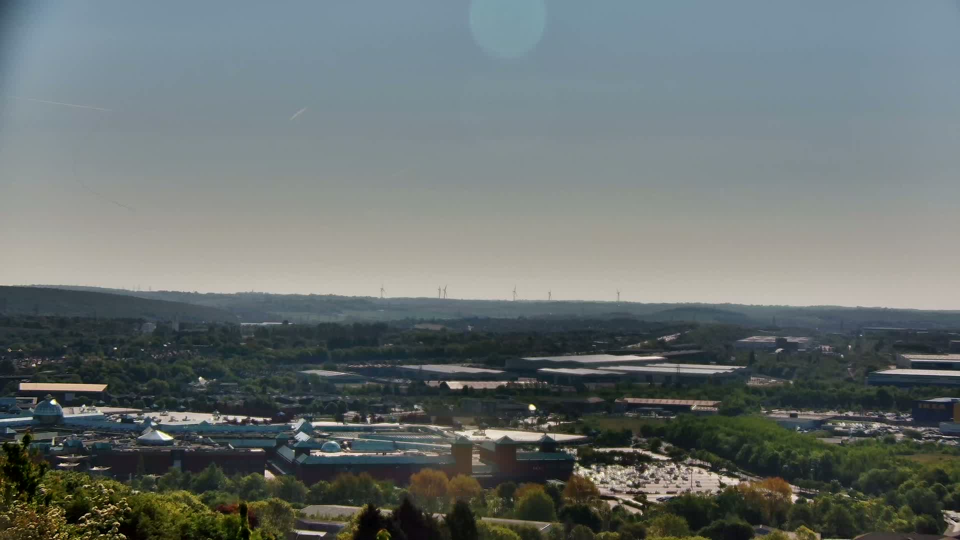 A sprawling suburban landscape with a large shopping center, industrial buildings, and a distant line of wind turbines under a clear, light-blue sky.